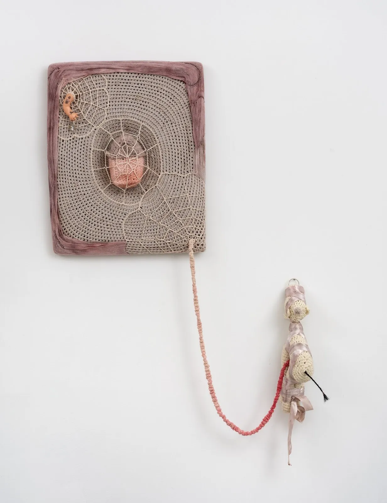 Wall-mounted textile sculpture with a web-like crochet pattern, a central pink object, and a dangling, wrapped appendage made of yarn and fabric.