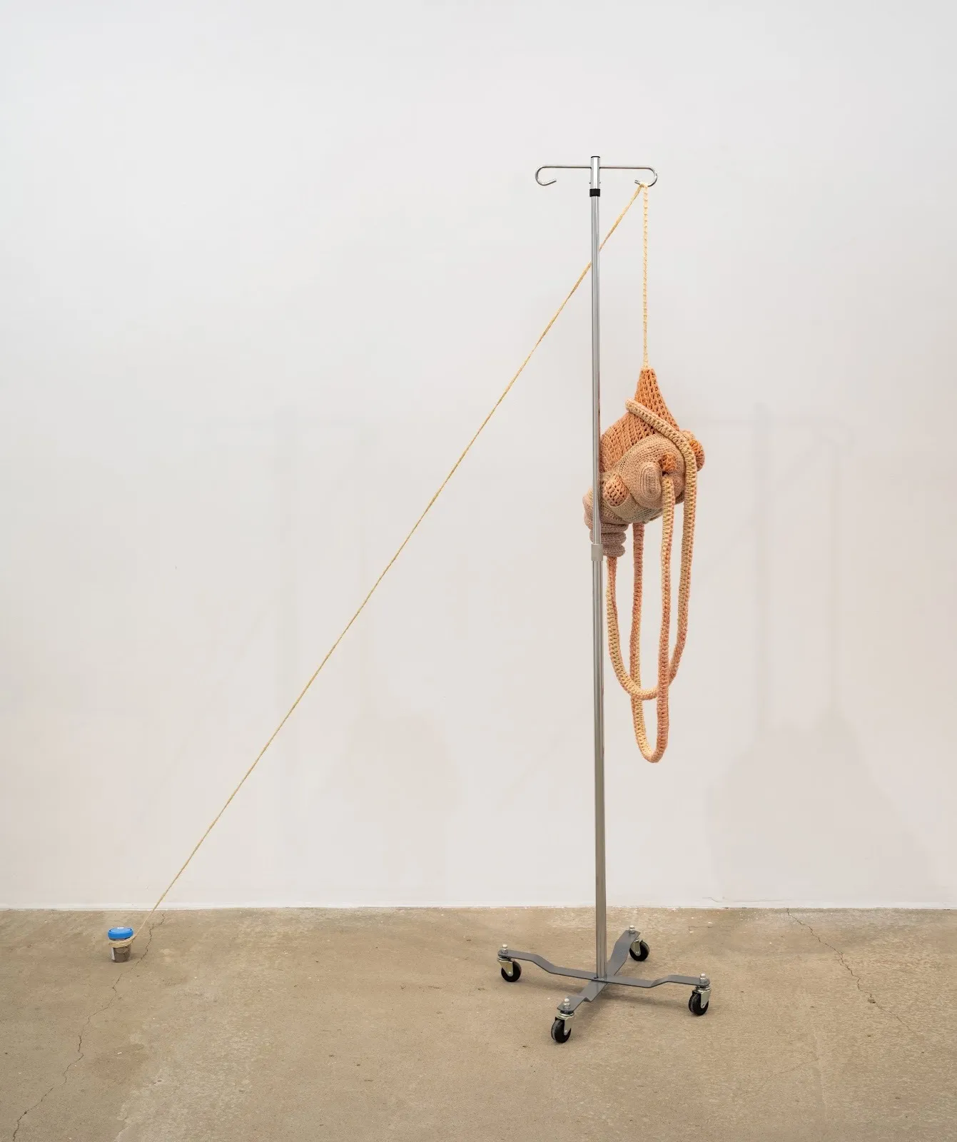 A soft, abstract textile sculpture in peach tones is suspended from an IV pole, with a long cord extending diagonally to a small object on the floor.