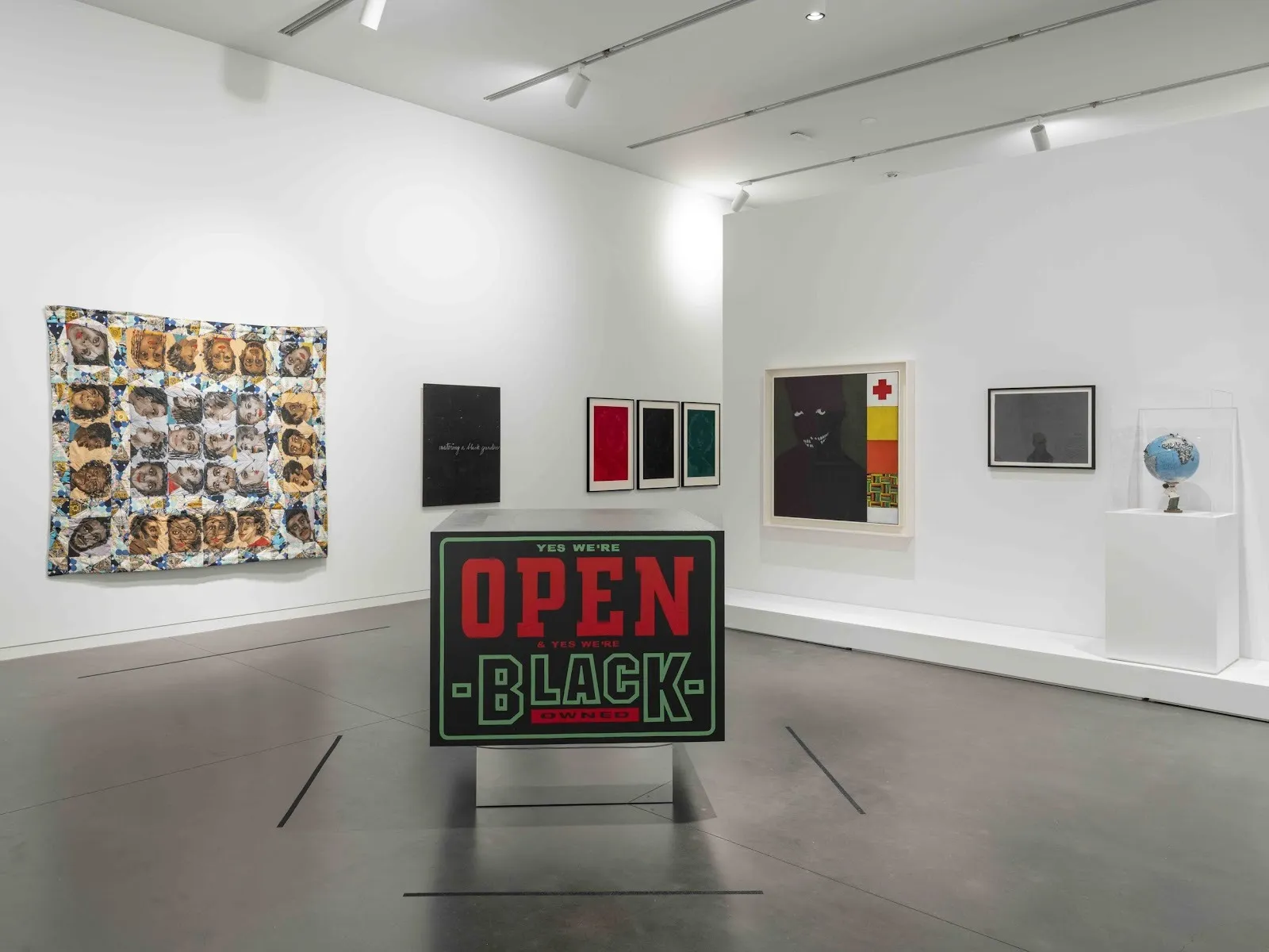 A large white gallery room holds multiple 2D and 3D works. The central 3D work rests on the concrete floor, a metal block with design text on the side that reads "YES WE'RE OPEN & YES WE'RE BLACK OWNED" in red and green. against black.