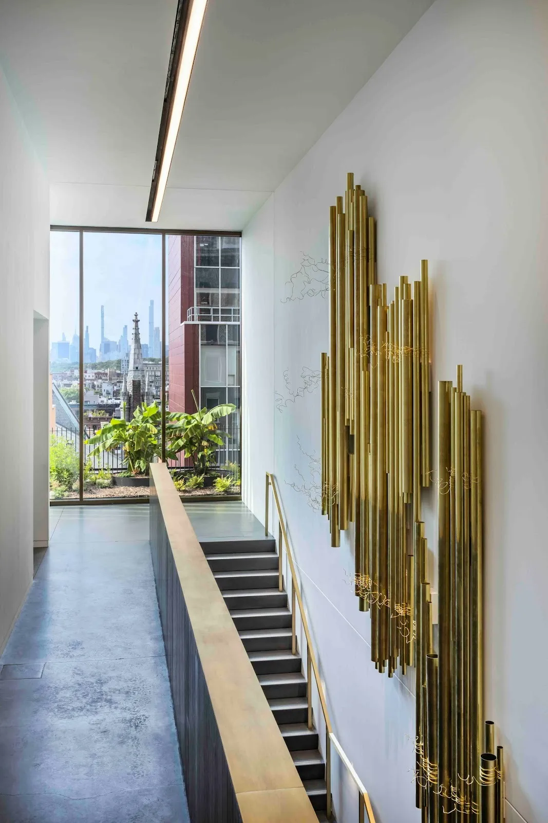 A spacious stairway with marble walls and floor-to-ceiling windows features a wall work on the side wall. The work is 3D, made from irregular groups of large gold tubes.