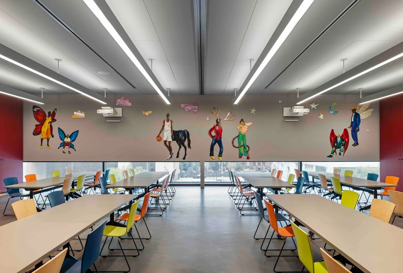 A large room with multiple long desks and colorful chairs features a mural on its far wall. The mural consists of human figures mixed with fantastical non-human parts, such as wings, horse legs, and tails.