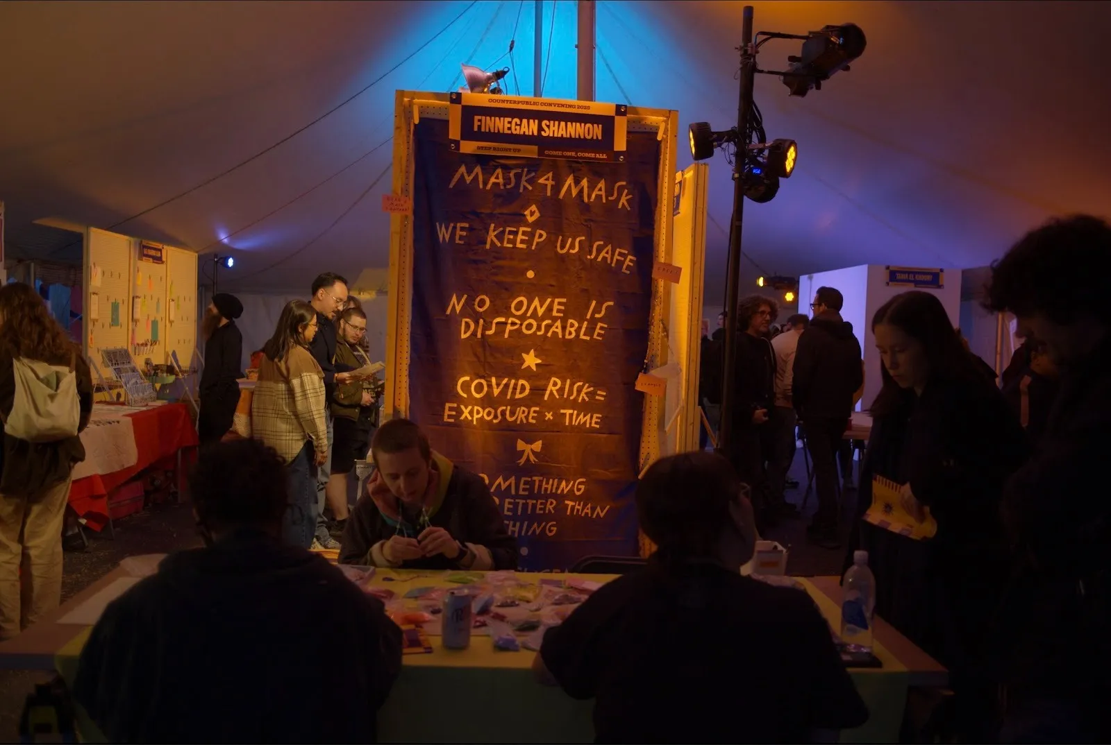 In the inside of a tent, people mill around table booths. The central table has a sign that reads "MASK4MASK, WE KEEP US SAFE, NO ONE IS DISPOSABLE."