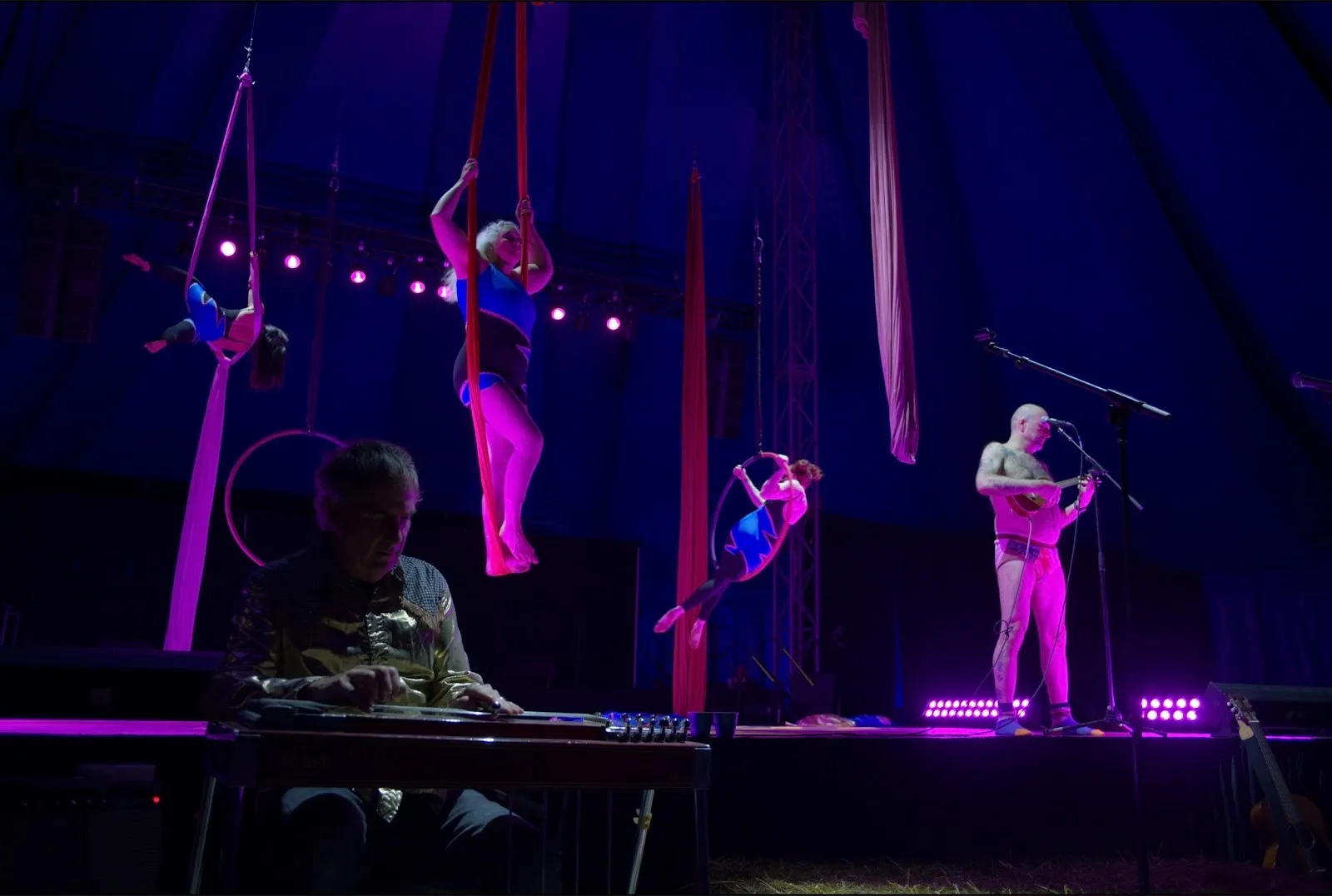 Inside a dimly blue and purple-lit tent, acrobats hang from aerial ropes and hoops while performers play instruments.