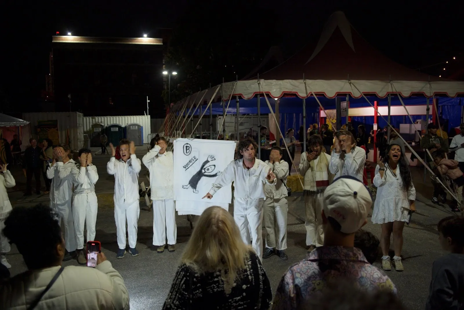 Outside a fair ground, people gather around a troupe of performers clad in white. They carry signage and enact gestures, their hands covering their face, or performing surprise.