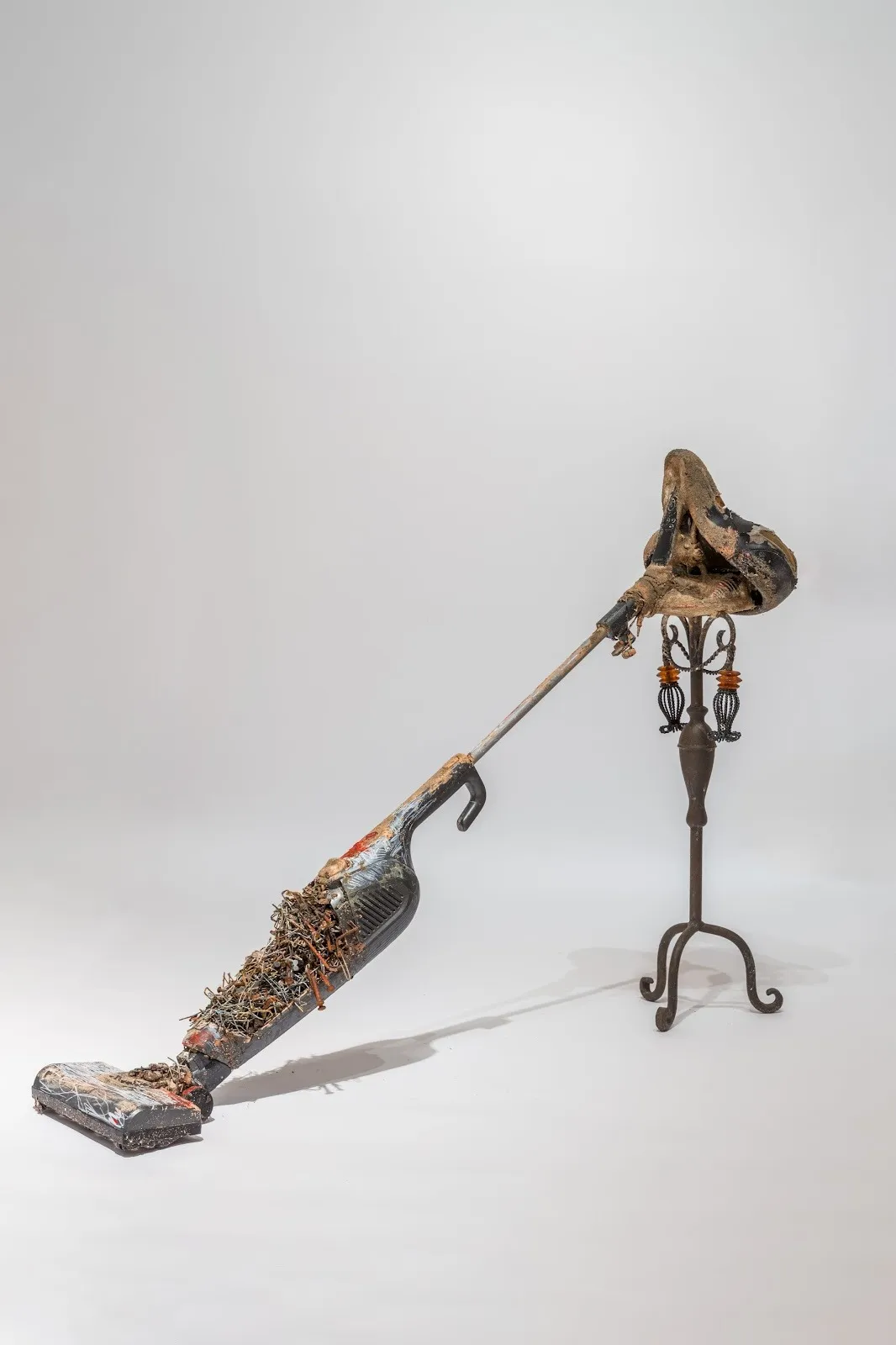 A sculpture made of amalgamated composite found objects features a worn vacuum covered in small metal pieces. The handle is a dirt-crusted bike seat, which leans on a small decorative tripod.