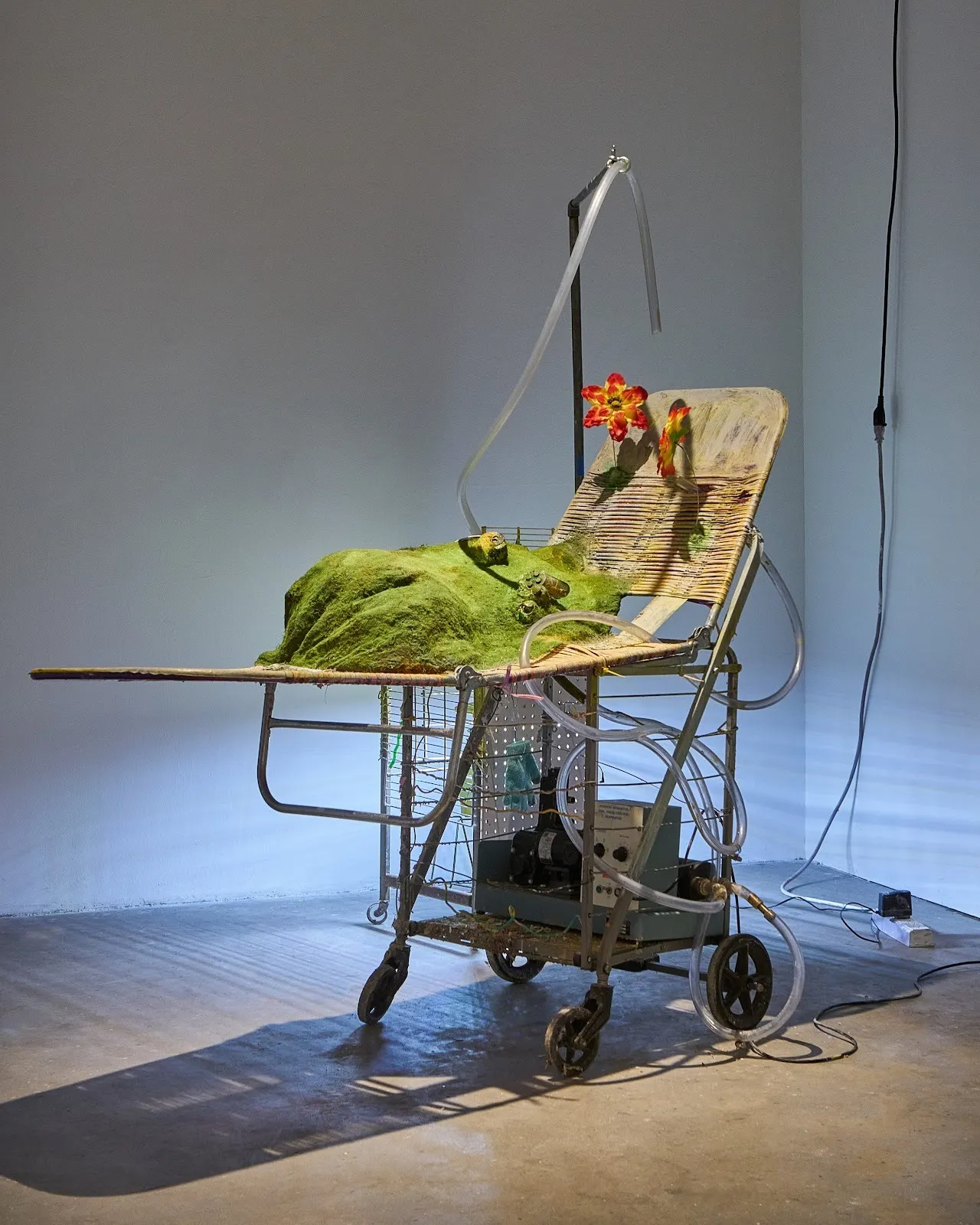 A woven lounge chair resting on a rolling cart is outfitted with flowers, flock grass, and a tubing apparatus that laces through the main structure and hangs on an adjoining pole stand.