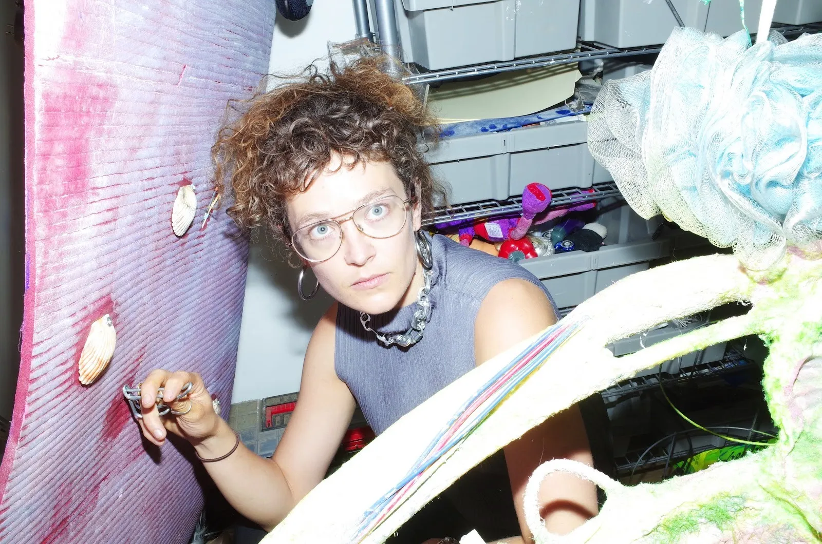 In a studio space crowded by bins, objects, and artworks, a woman looks above the camera, crouching next to a pink surface with shells, holding a metal tool to the surface.