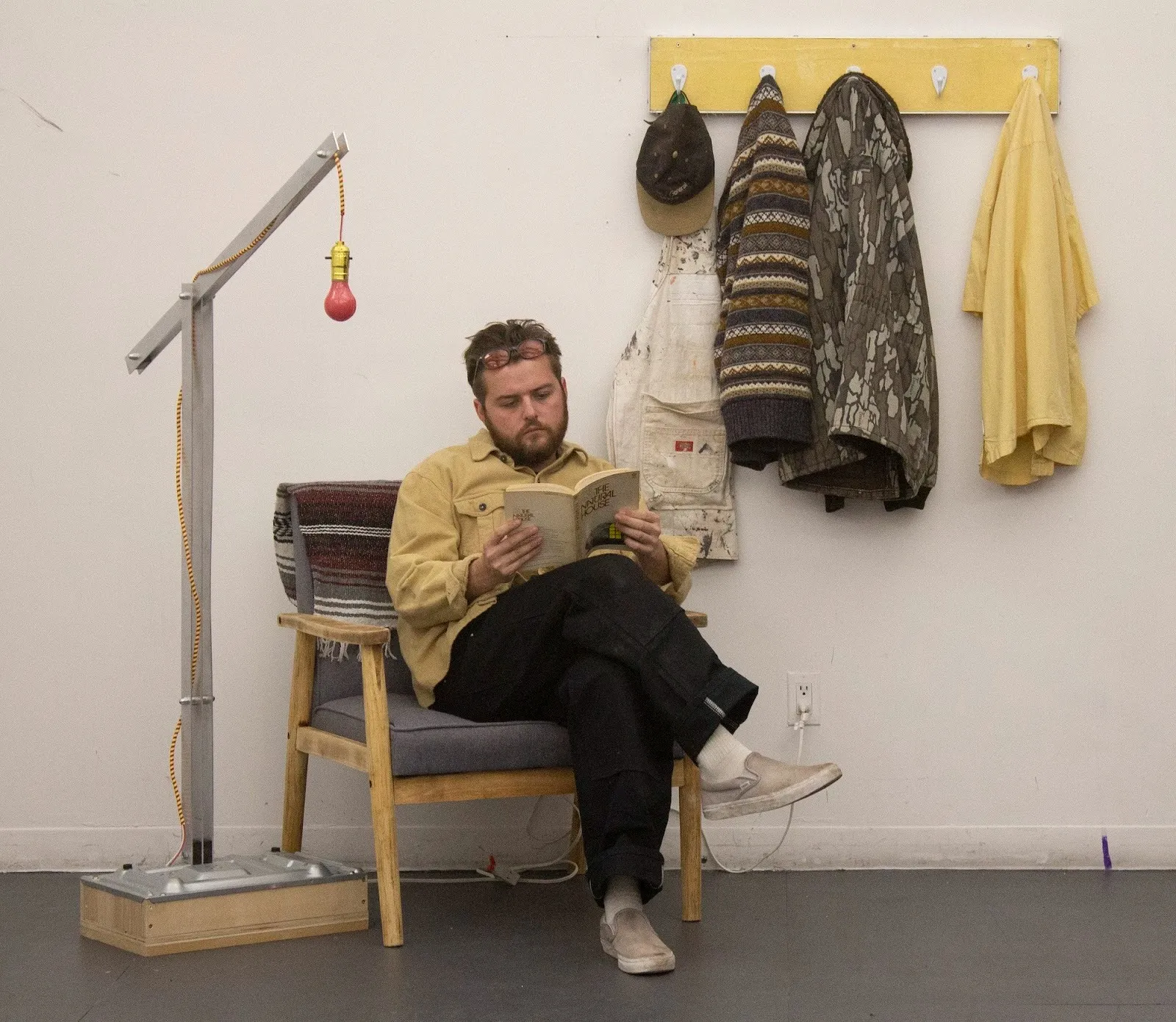 A man sits in a wooden chair reading a book under a red hanging lightbulb, with coats and clothing hanging on a wall-mounted rack behind him.