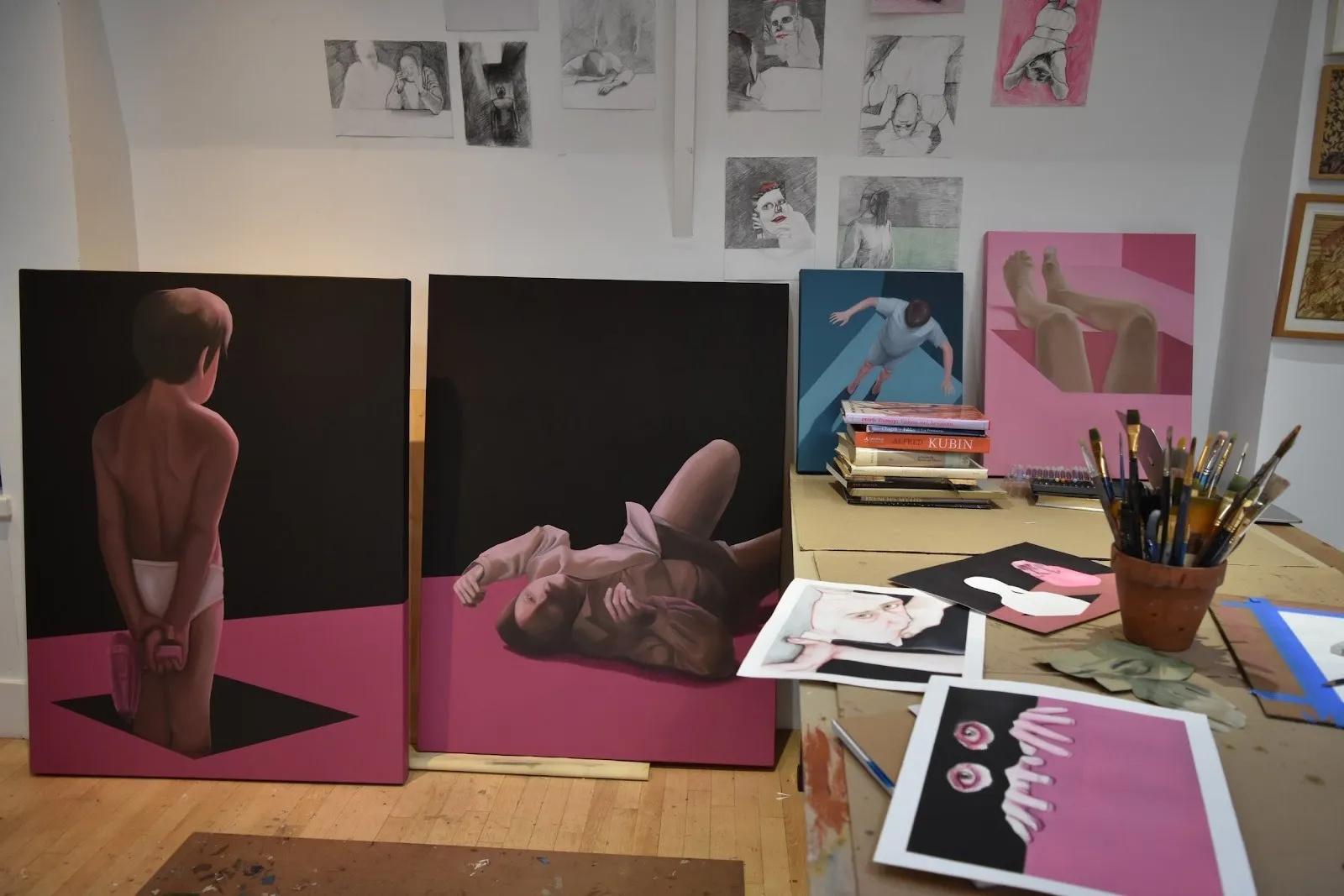 A workspace filled with surreal, figurative paintings on black and pink backgrounds, surrounded by sketches on the wall, paintbrushes, and art books on a table.