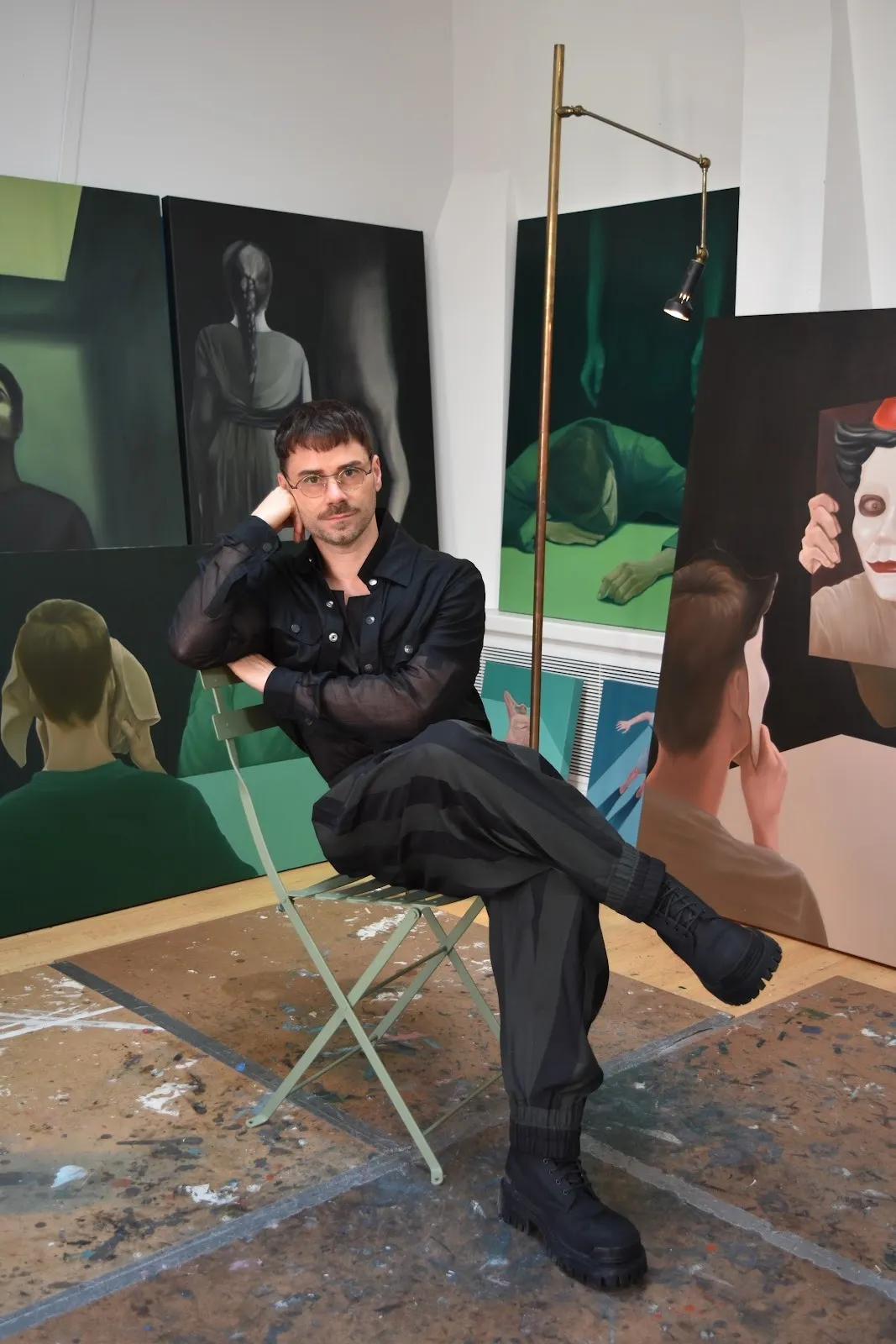 A male artist in dark clothing sits on a green chair in a studio, surrounded by moody, surreal paintings featuring partially obscured figures in dramatic lighting.