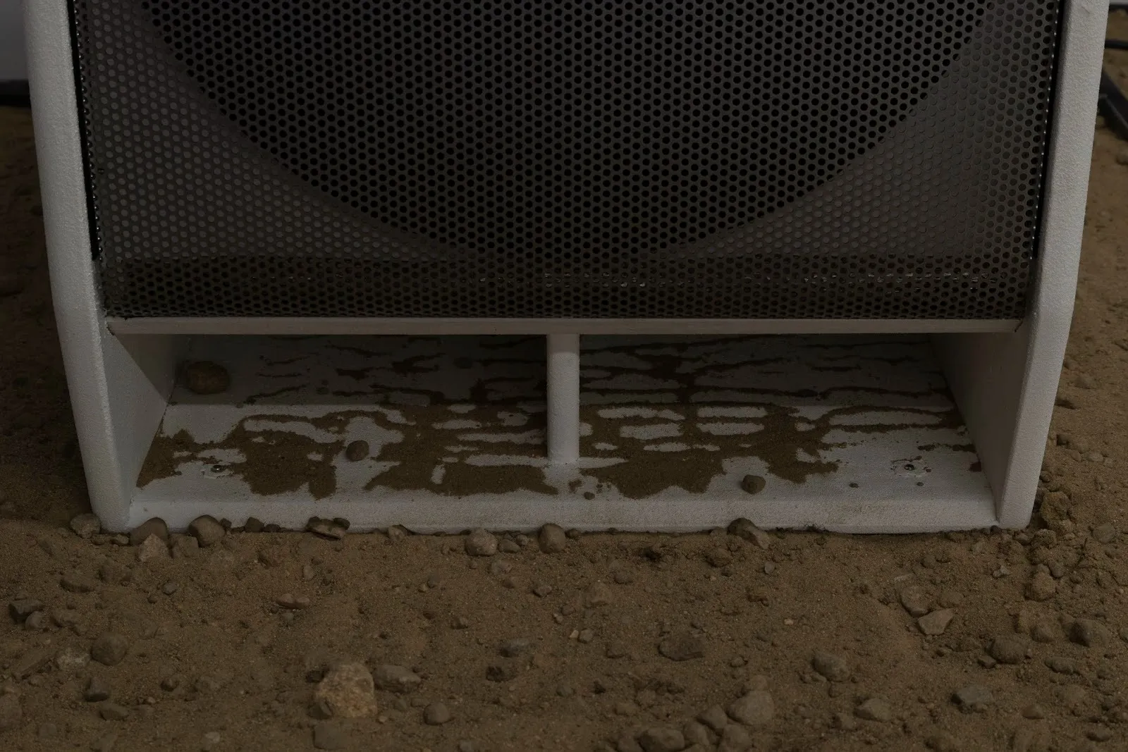 A speaker resting on a pebbled dirt surface features two compartments below the speaker's screened mouth. Each compartment holds lines of sand, shaped by the speaker's vibrations.