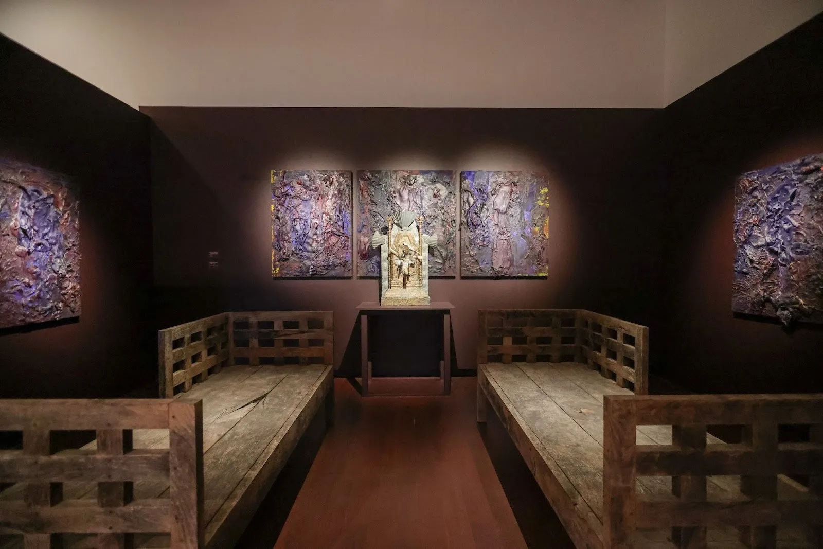 Intimate chapel-like gallery with purple-toned paintings, a central shrine, and two rustic wooden benches facing each other.