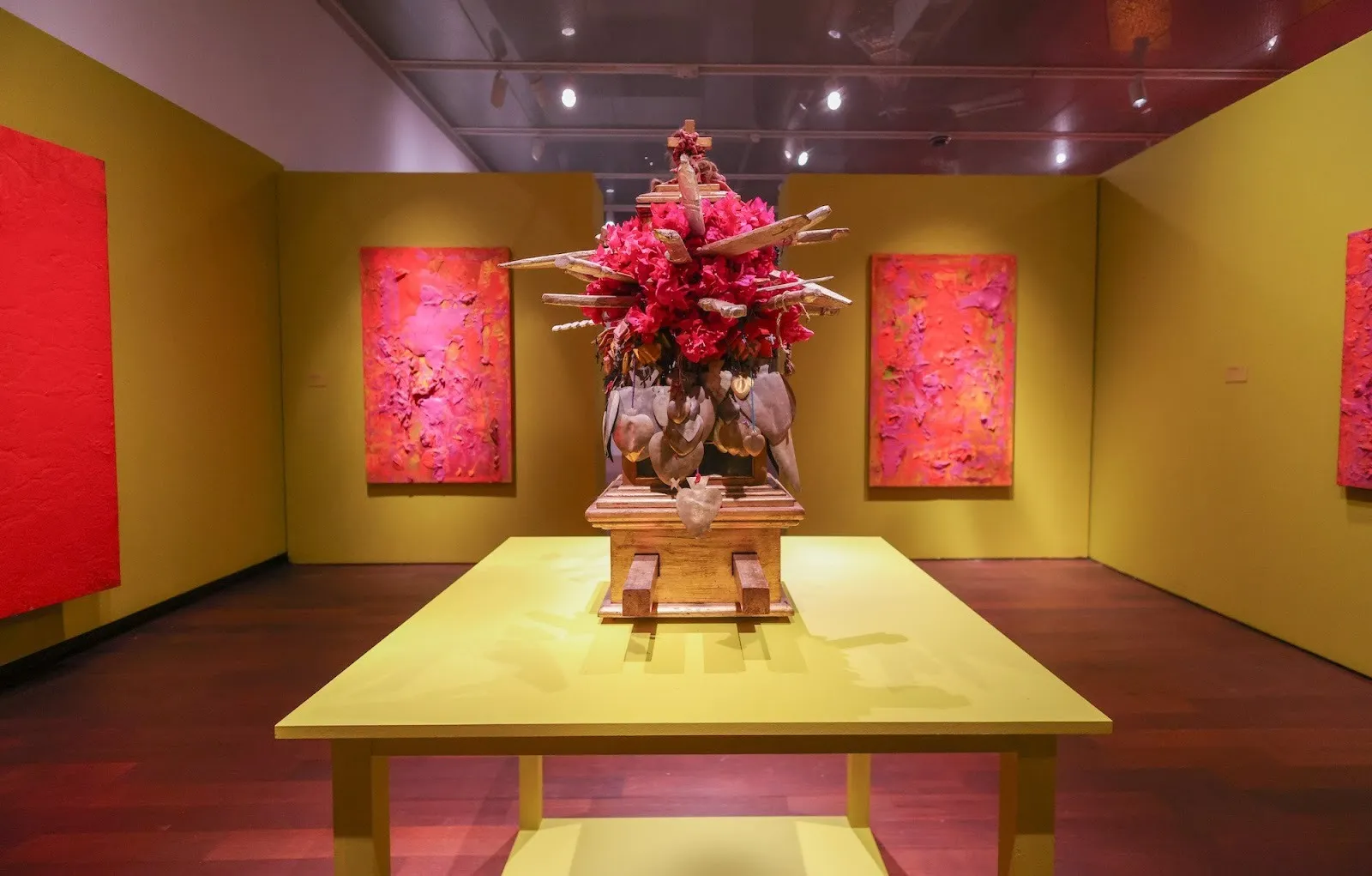 Mixed-media shrine adorned with red flowers and sacred heart votives, centered in a bright yellow room with textured pink paintings on the walls.