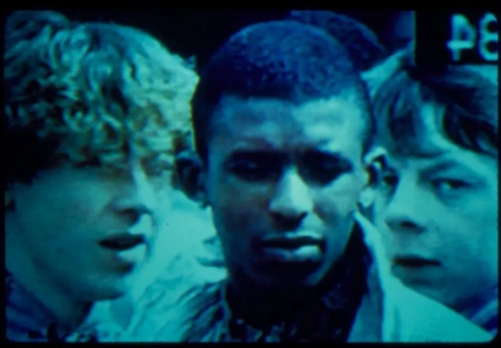 A grainy, blue-tinted still of three young men closely grouped together, facing the camera.