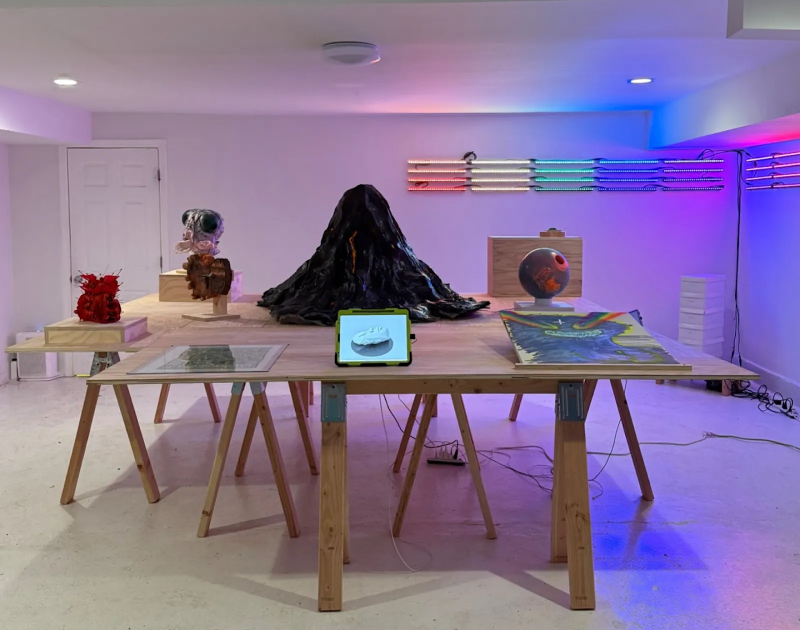 In a neon-lit room, a wooden desk holds various art objects including a miniature mountain, a globe, and an ipad, report from nori mountain on view at smilers, photo by laura tighe.