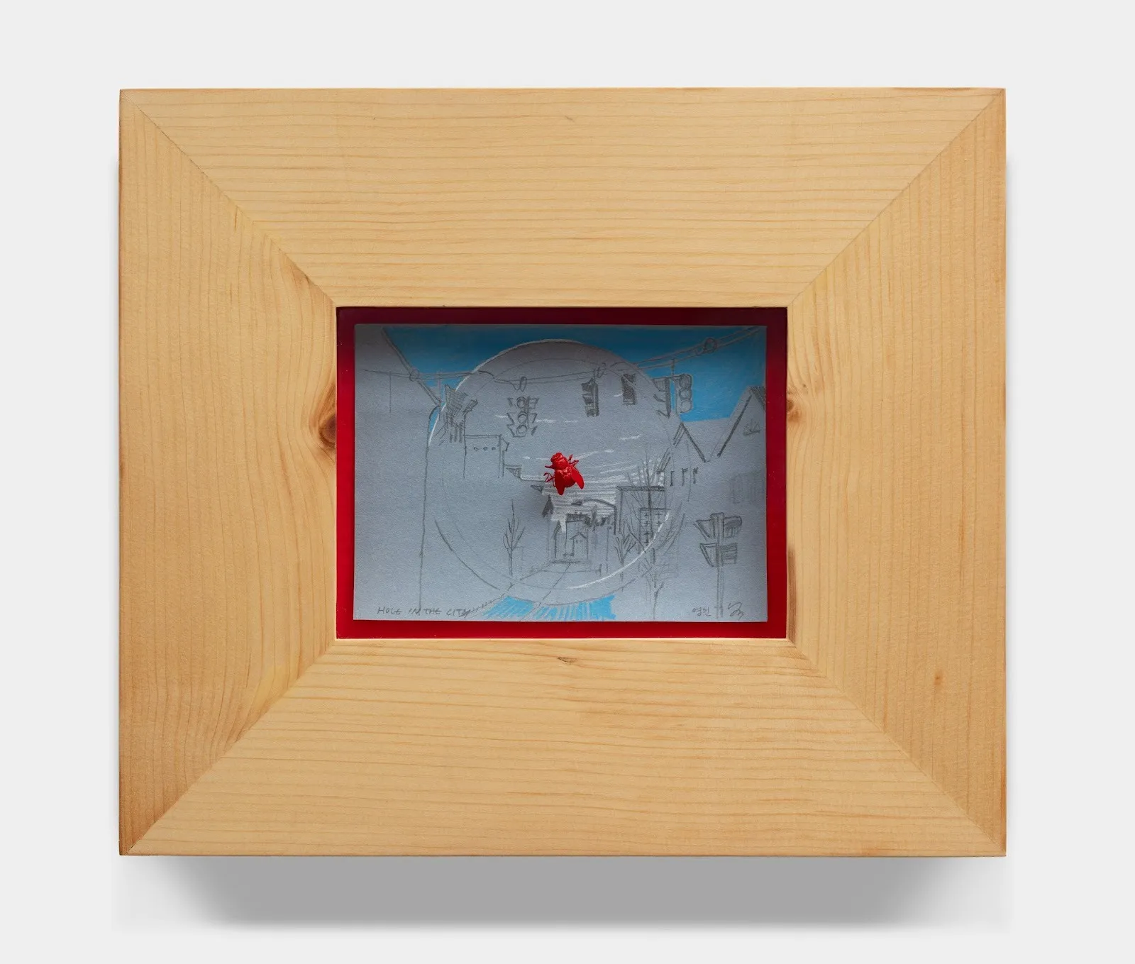 A painting is framed by thick, raw wood. Against a thin red lining, a small sketch of a cityscape done in grey and blue rests. In the center, a red ceramic fly is pinned to the work.