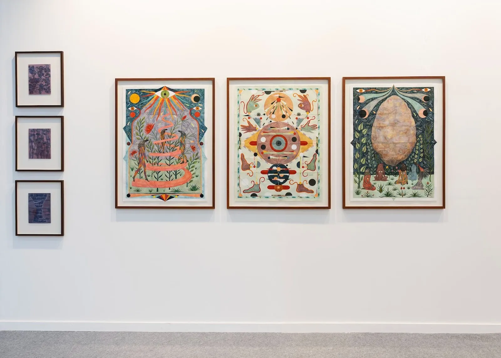 A gallery wall displaying a row of framed works, including three large colorful, symbolic illustrations flanked by smaller abstract studies.