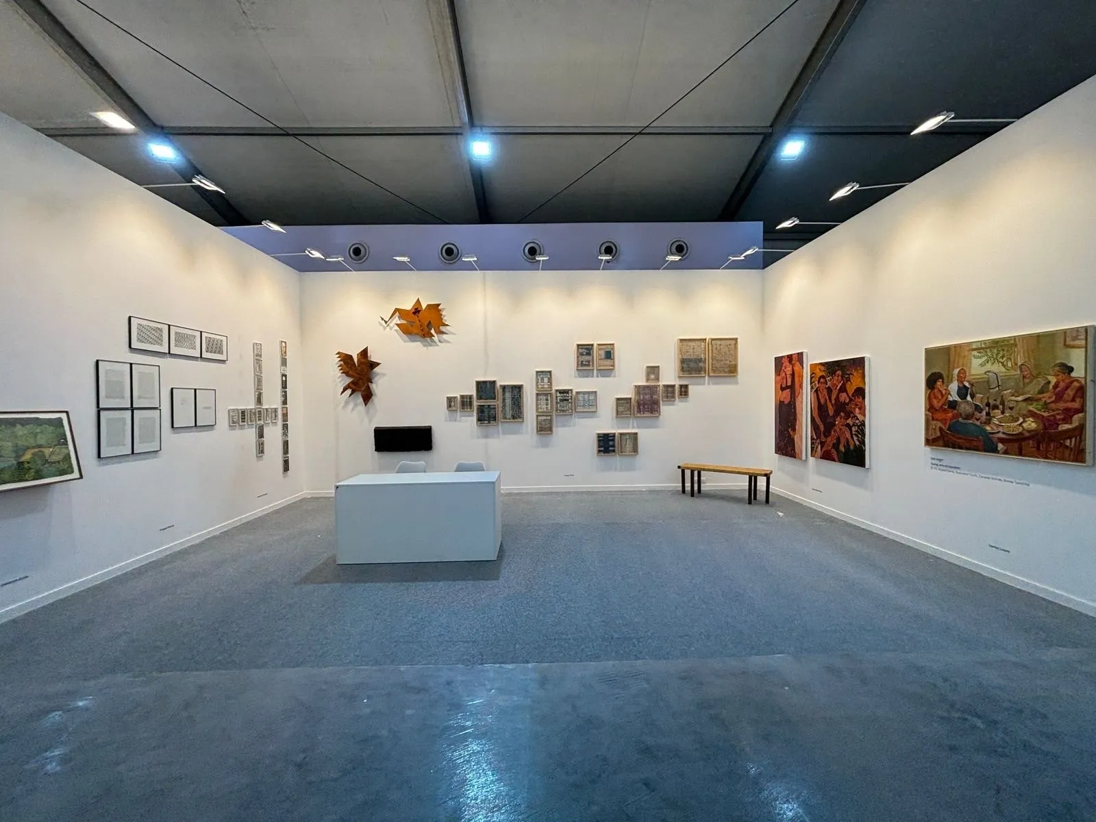 A spacious gallery booth with a wide range of framed artworks, wall-mounted sculptures, and a central bench under bright overhead lighting.