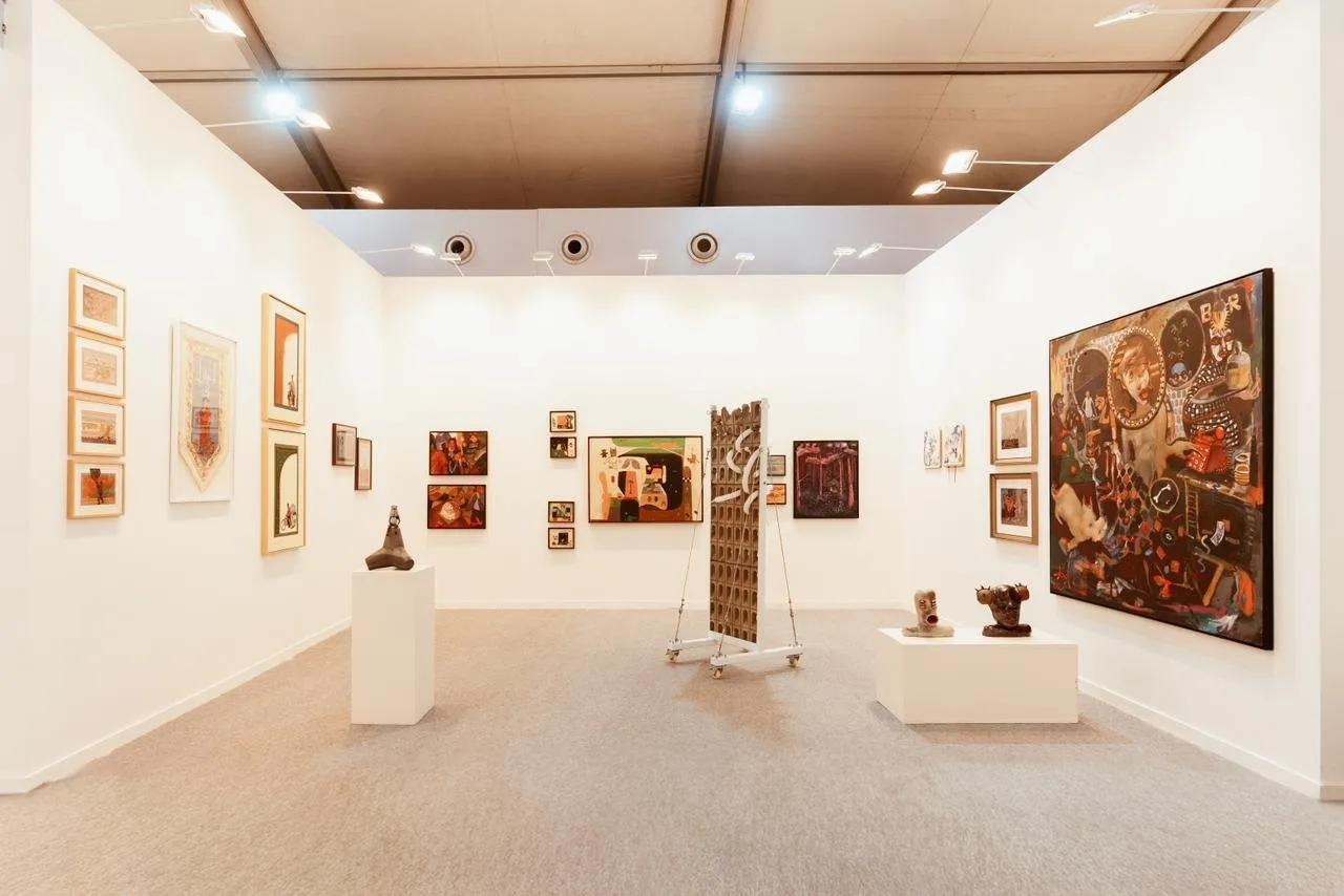 A gallery booth displaying an eclectic mix of paintings, framed works, and sculptures, with a freestanding sculptural screen in the center.