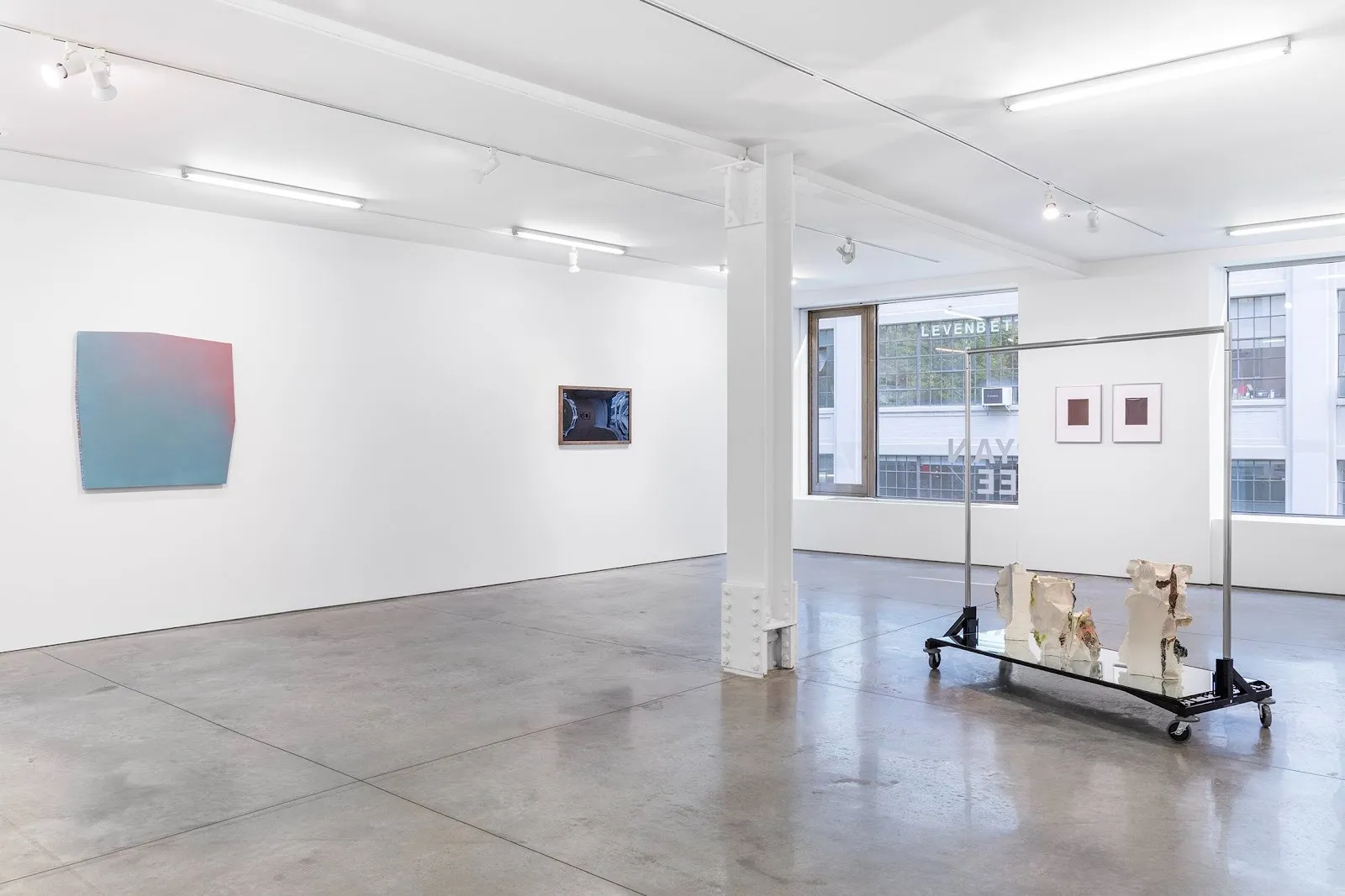 A spacious art gallery with minimalistic white walls, showcasing abstract artworks. On the left, a soft gradient painting hangs, and in the center, a sculpture on a rolling cart is displayed next to framed prints. Large windows let in natural light.