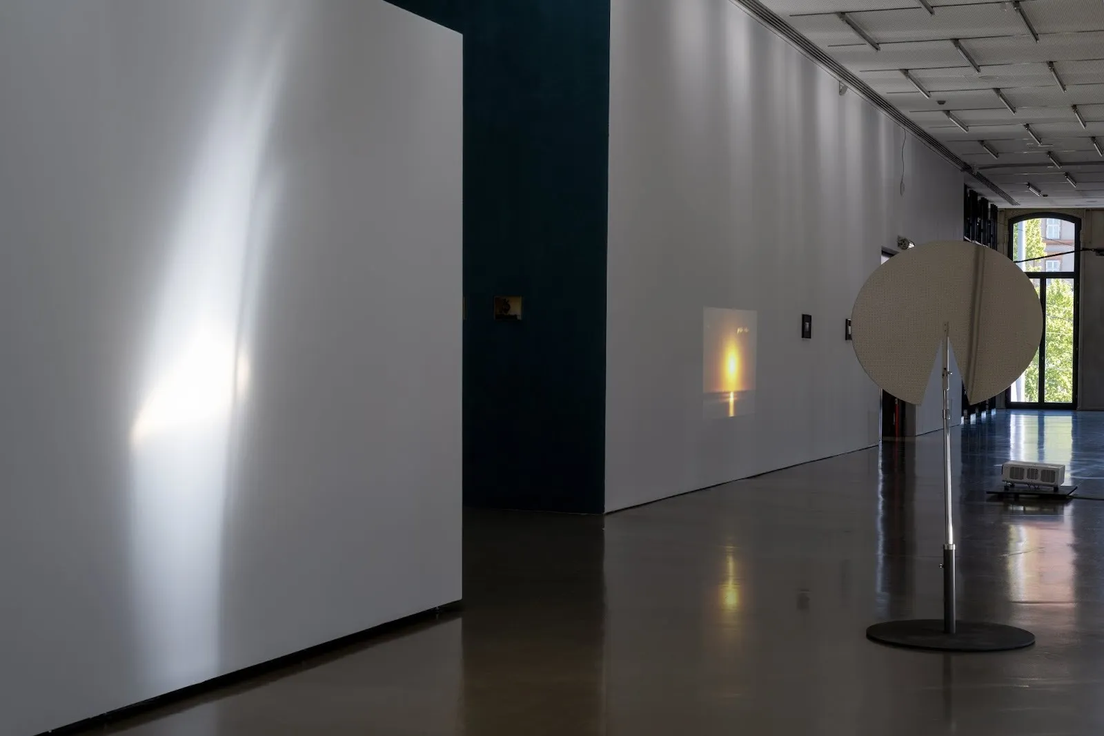 A long gallery corridor with a faint beam of light on a white wall, a small warm-toned projection ahead, and a large circular metal sculpture placed on the right.