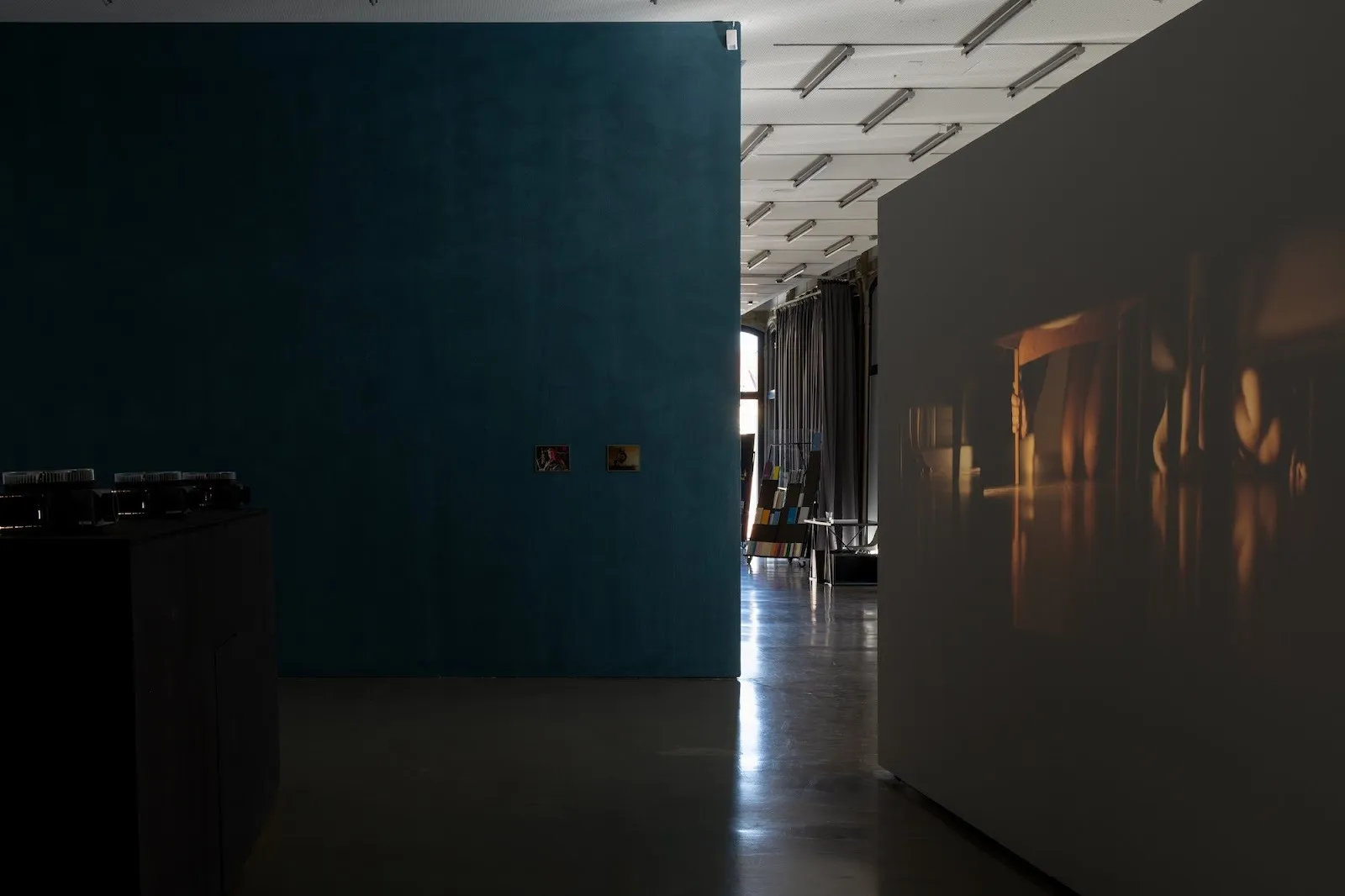 A dim gallery corner where a warm-toned projection of feet under a table appears on a wall, with two small artworks mounted on a teal-colored wall nearby.