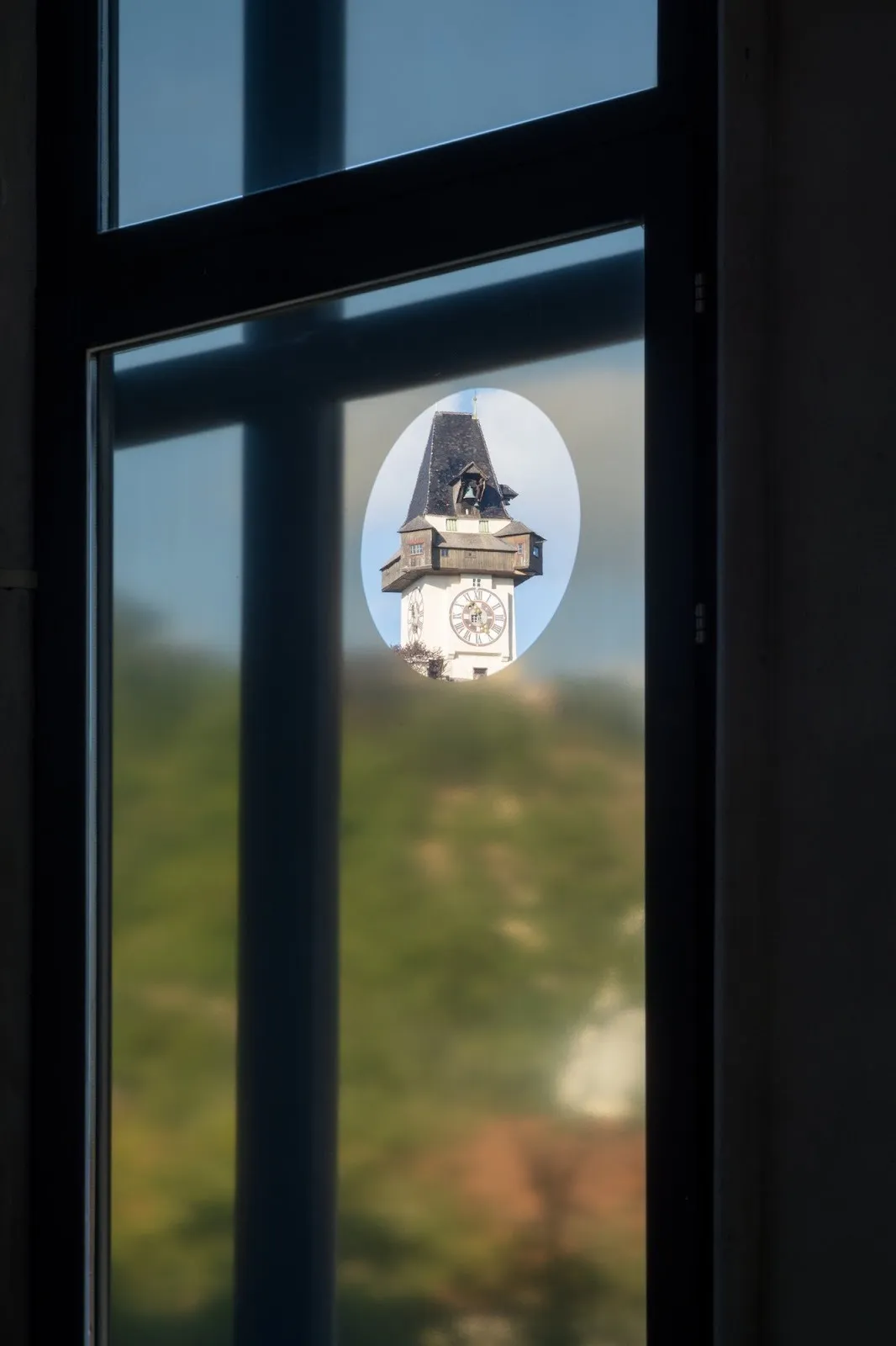 A view through a frosted window revealing a crisp oval opening that frames a historic clock tower against a blue sky.