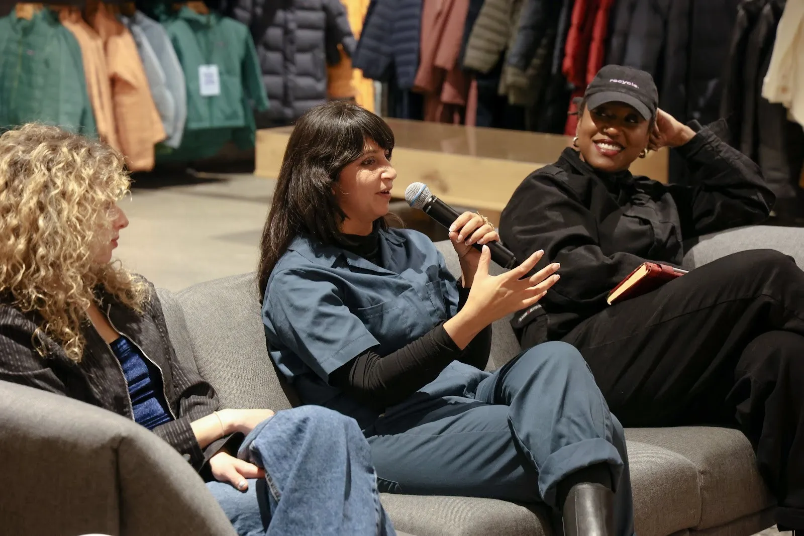 Three people sit on a grey couch and a large clothing store. The middle person, who wears blue coveralls, gestures while speaking to an audience into a microphone.