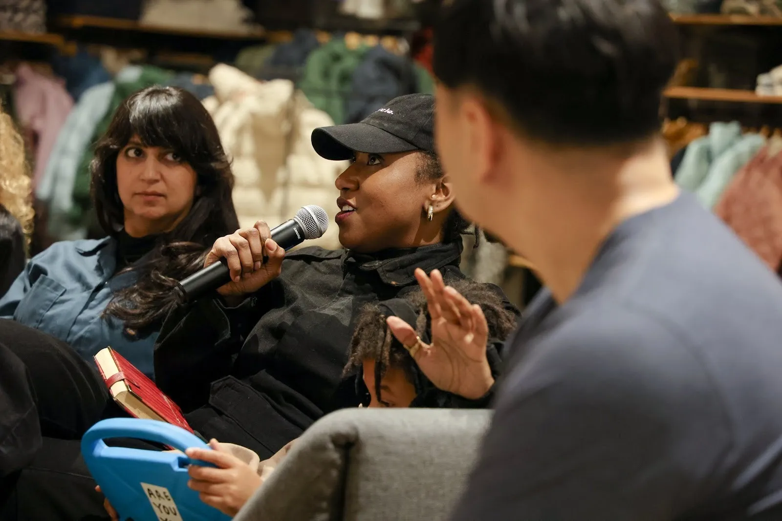 A person wearing a black cap sits on a grey couch and speaks into a microphone. One person on the right and one person on the left listen to them.