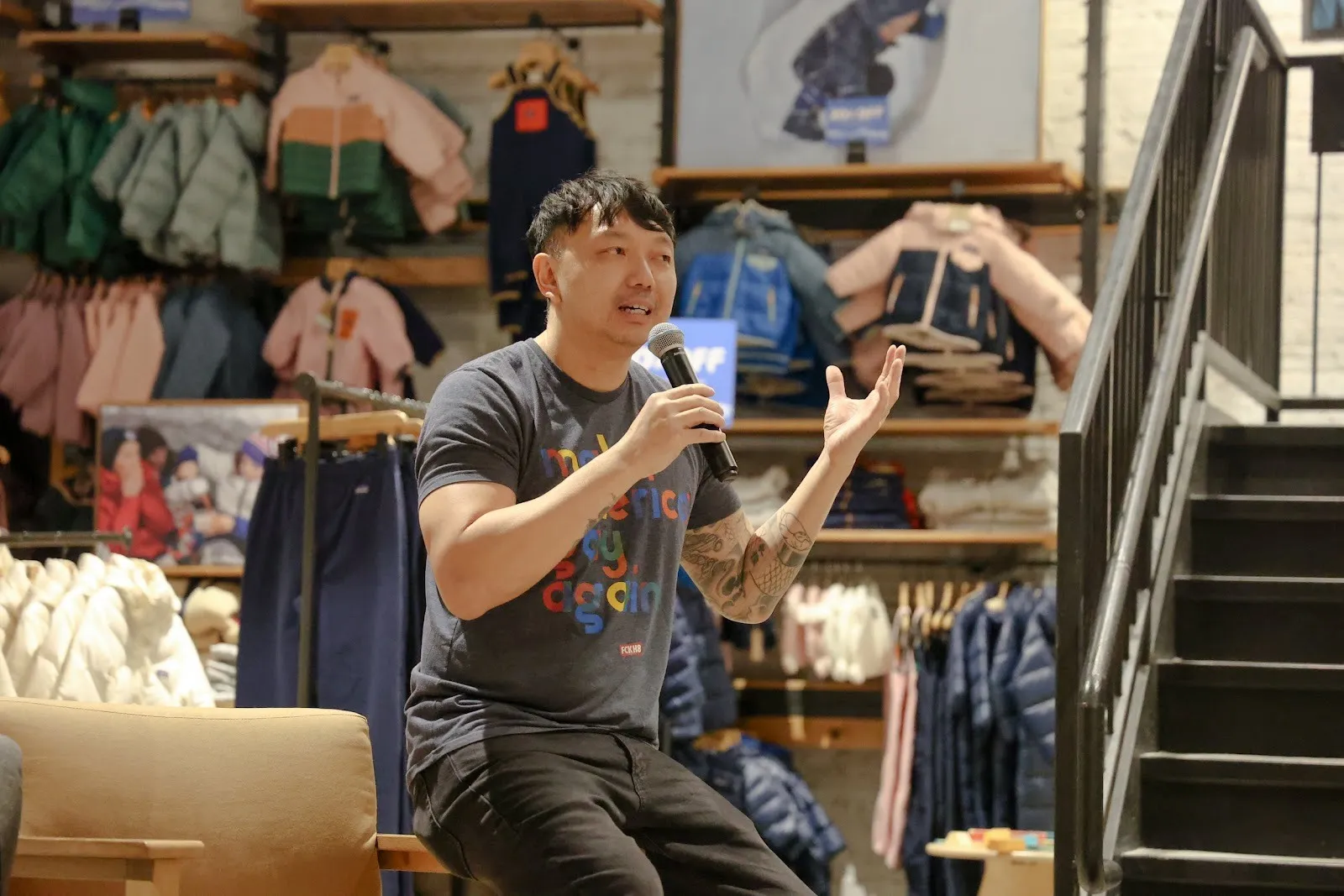 In a clothing store, a man in a grey and lettered t-shirt sits on the armrest of a chair, speaking into a microphone and gesturing.