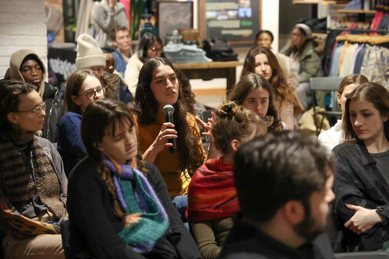 In a seated indoor audience, a person wearing a yellow sweater speaks into a microphone, asking a question.