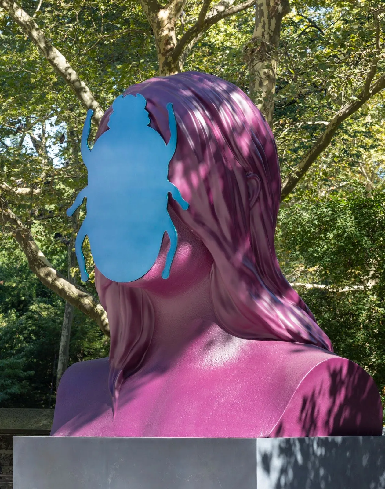 Close-up of the magenta bust sculpture showing the smooth, stylized hair and a metallic blue beetle silhouette obscuring the face, dappled with sunlight from overhead trees.