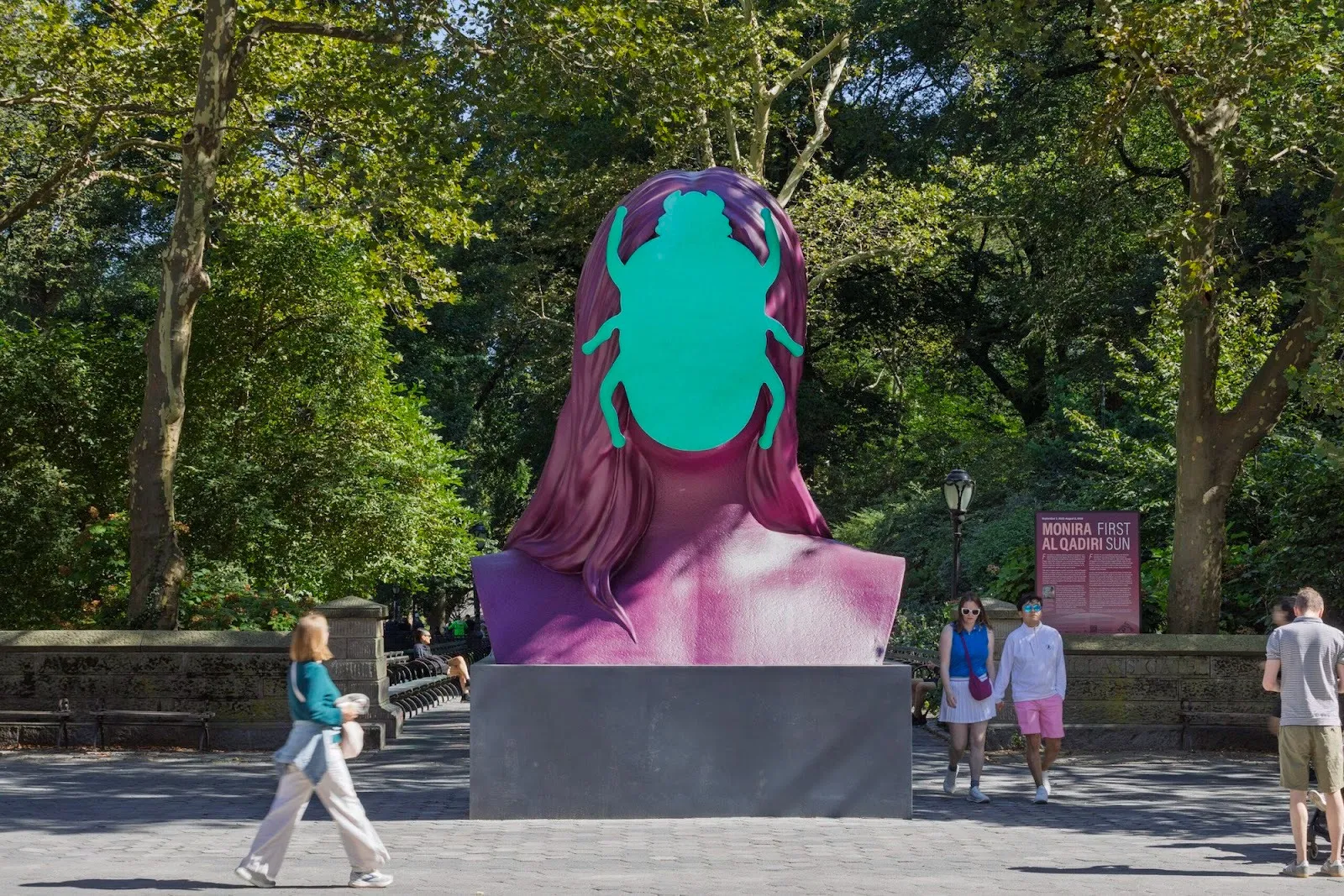 A large magenta bust sculpture with long hair and a flat, bright green beetle shape covering the face, installed outdoors in a tree-lined plaza with people walking by.