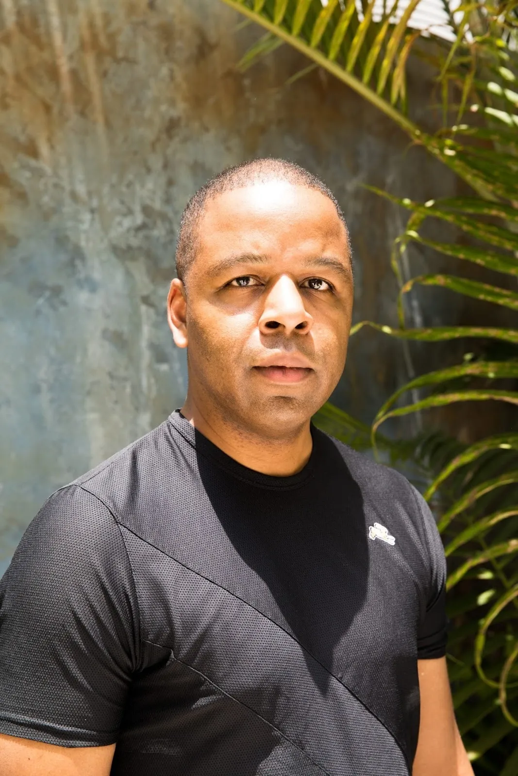 A man with closely shaved hair and a black athletic t-shirt stares at the camera, with plants and concrete behind him.