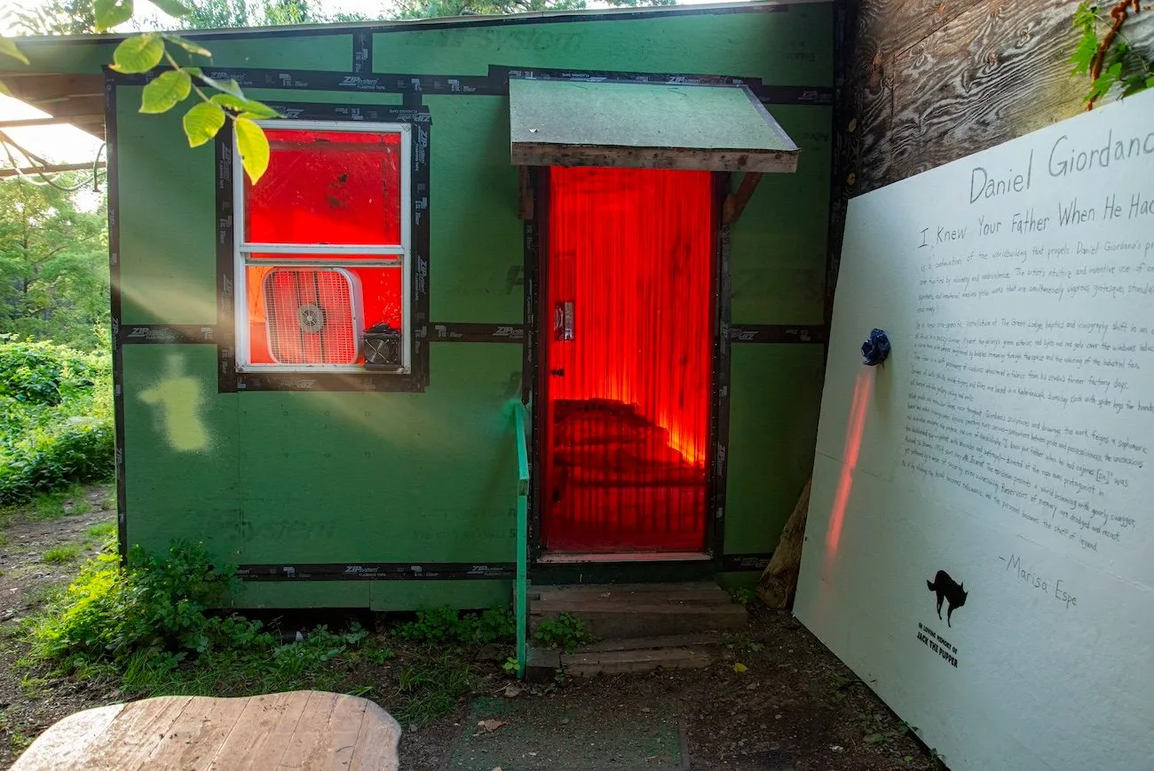 A green shed-like structure in a foresty space is lit with red light from within. To the side of the shed, a painted white wooden board leans against exposed wood and features handwritten text.