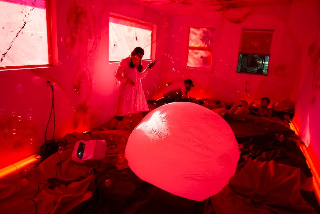 In a room lit with red light, people stand and lie down on its floor, which is covered in mattresses and sheets. An inflated ball-like structure dominates the room and bubbles float throughout.