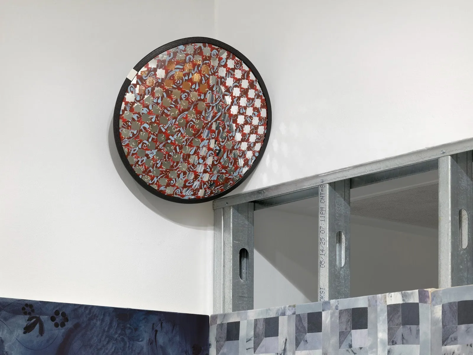 Detail of a gallery corner. Above exposed steel beams hangs a convex mirror painted with red and blue strokes that reveal star-shaped patterns of exposed mirror. At the bottom of the image, the edges of a painting peek into the frame.