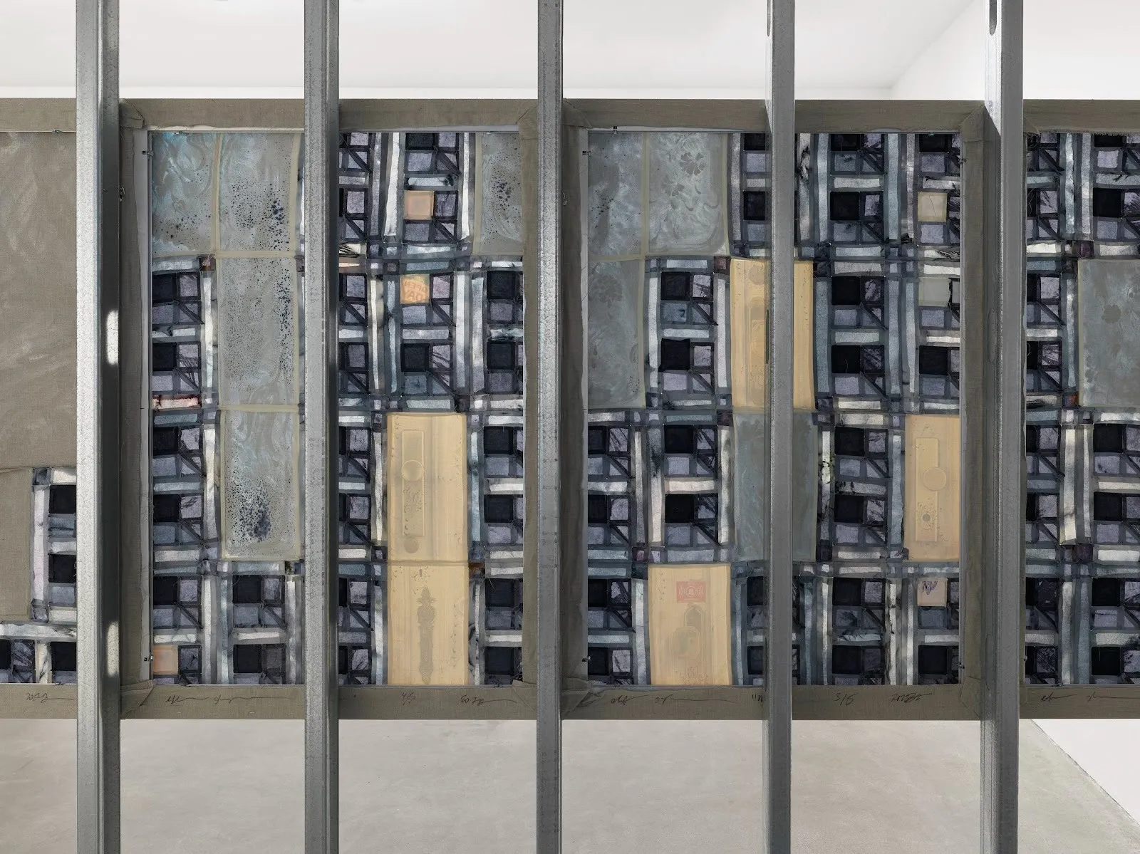 A view of the back side of a painting suspended across five steel beams. The back features a blue patchwork pattern with beige rectangles dispersed throughout. Faint outlines of doorknobs are visible through some of the beige rectangles.