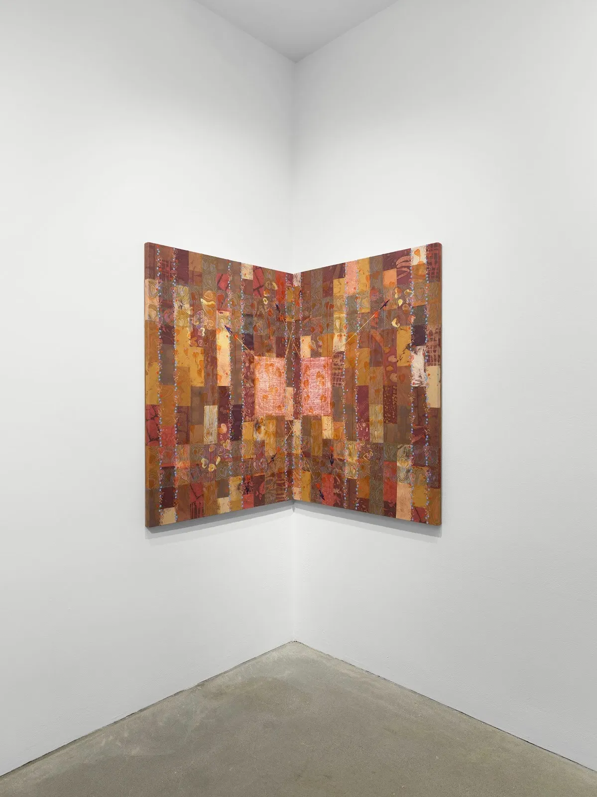 Painting featuring a brick-like pattern in shades of red, brown, pink, and orange is positioned in the corner of a white-walled gallery. The brick patterns have a wood-grain like tecture to them, with heart and floral motifs scattered throughout.