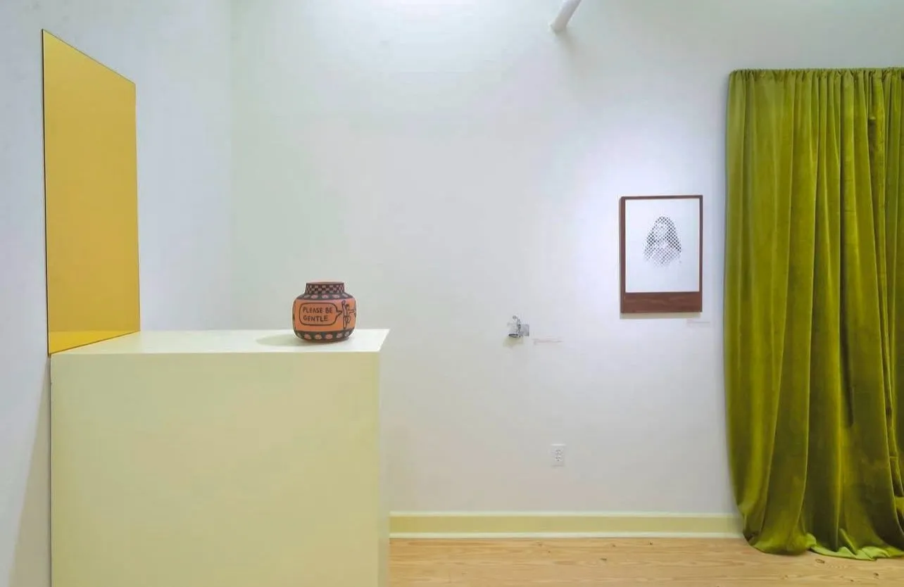 Exhibition view with a ceramic pot reading “PLEASE BE GENTLE” on a pedestal, yellow wall panel, framed portrait cut-out, and green curtain.