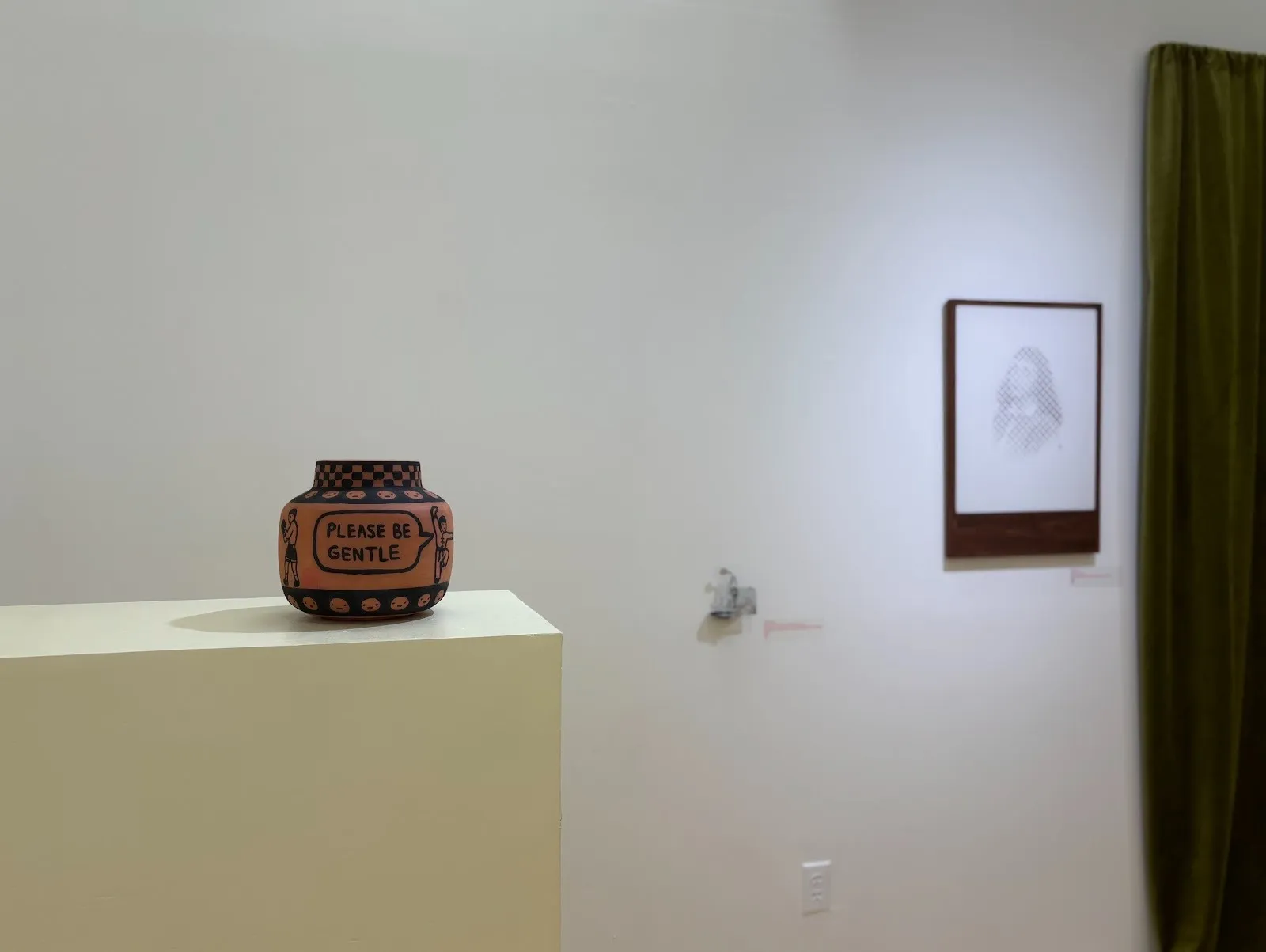 Ceramic pot on a pedestal with the text “PLEASE BE GENTLE,” displayed near a framed artwork and green curtain.