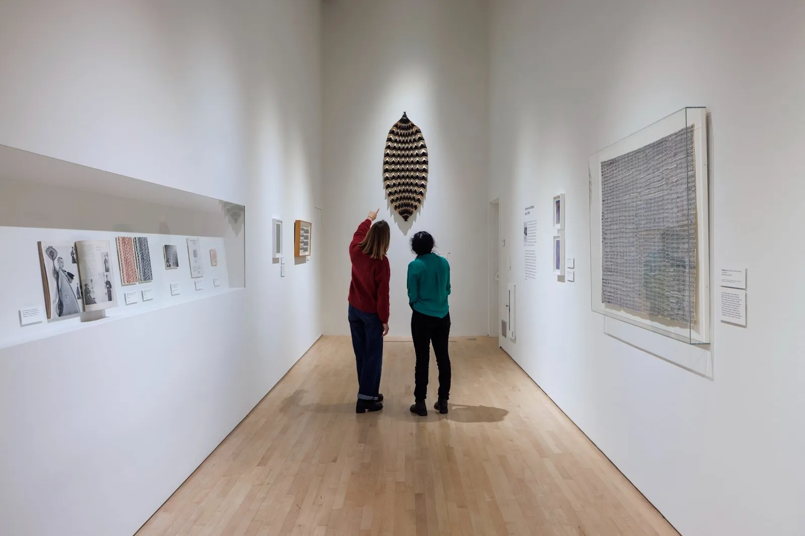 In a white museum corridor, two people look and point at a leaf-shaped woven sculpture hanging central on the far wall. Along the corridor walls, different 2-D works hang on walls and in vitrines build into the wall.