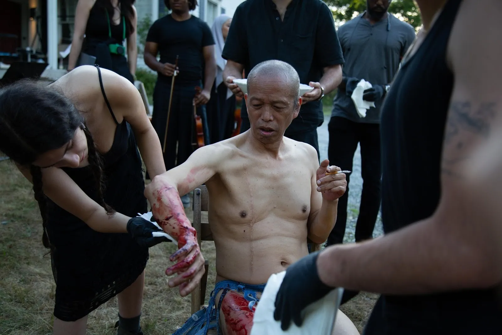 A shirtless man sits while others tend to his arm covered in blood, surrounded by musicians dressed in black.