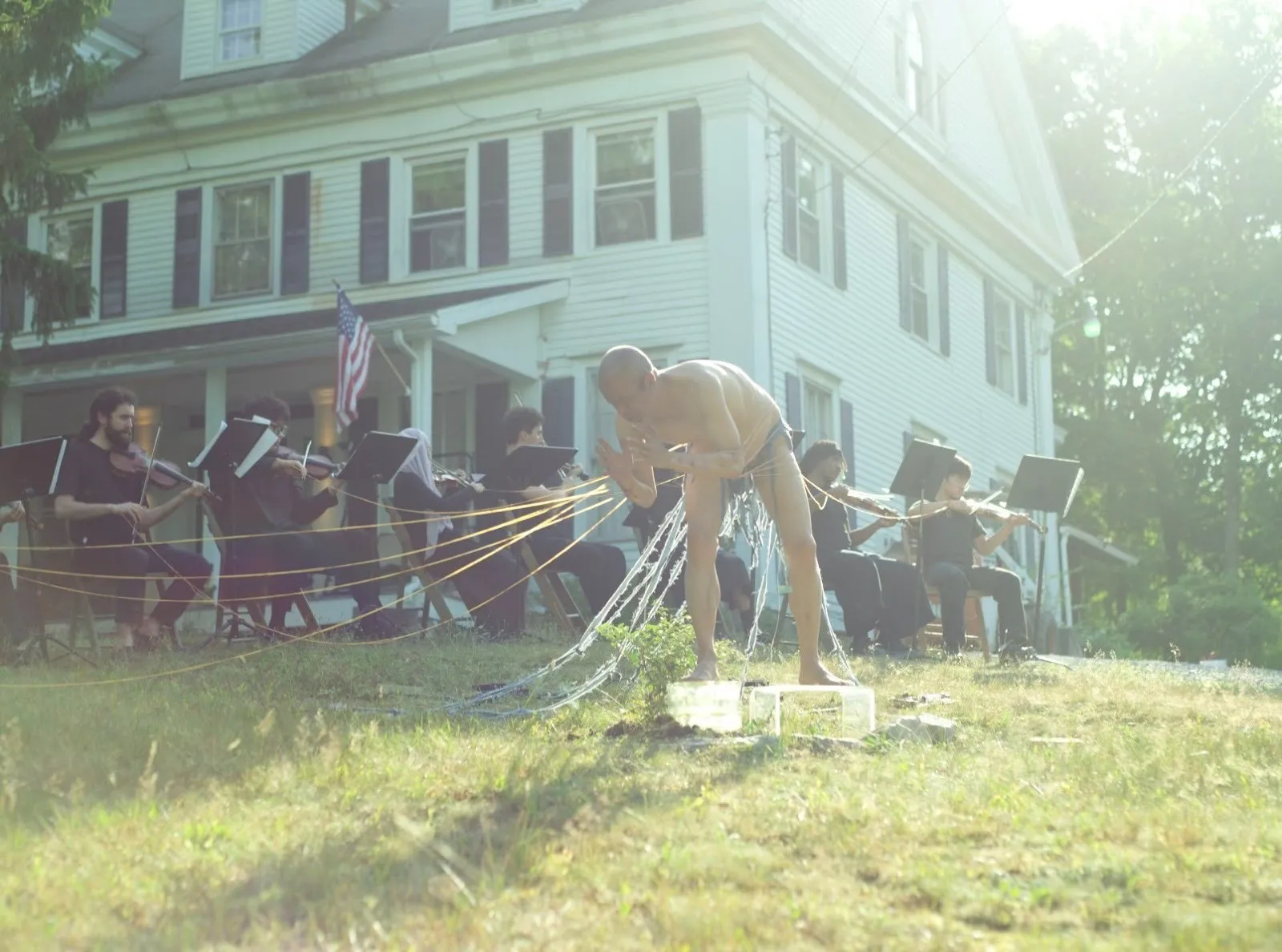A shirtless performer stands outdoors in front of a white house, connected by strings to a seated orchestra playing violins.
