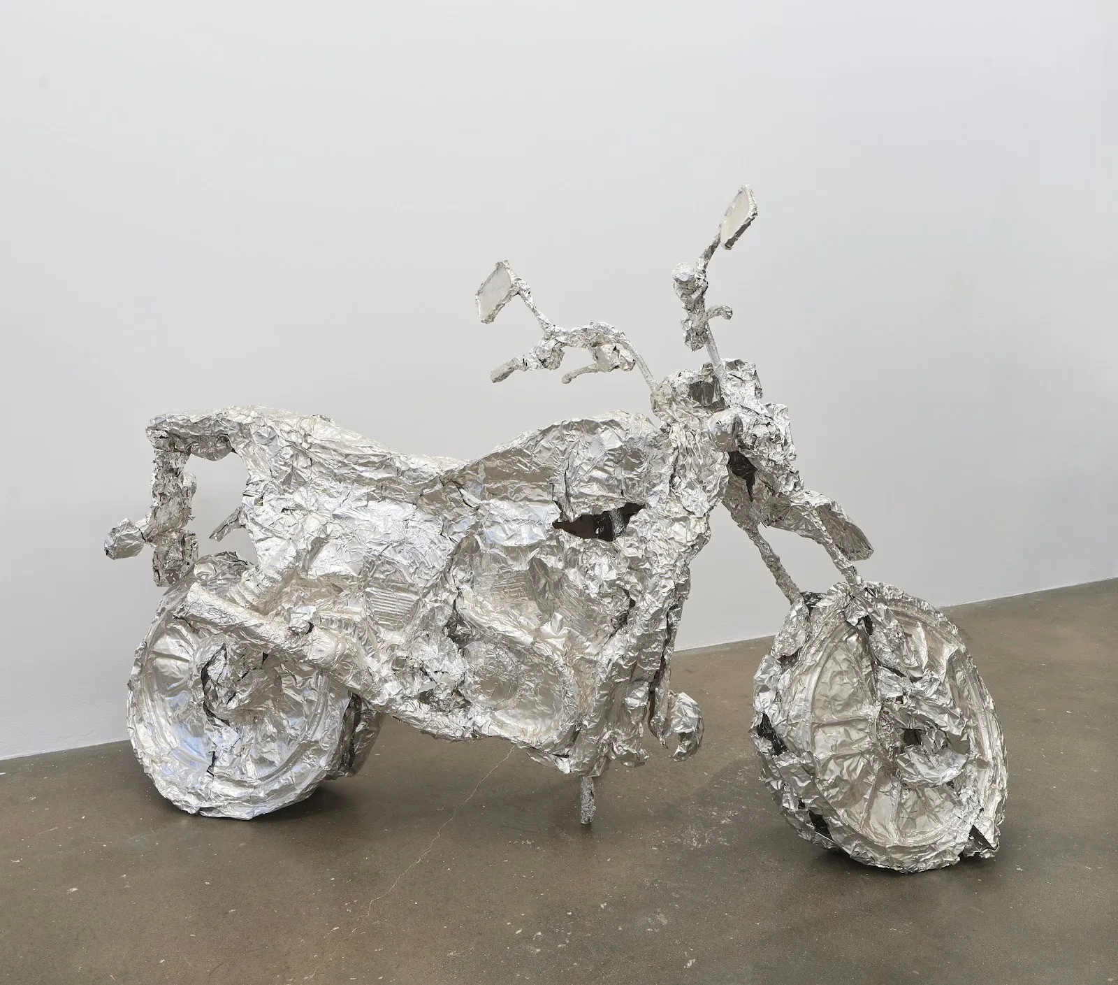 A motorcycle-like sculpture made of crumpled and molded aluminum foil sheets sits in a In a white gallery room.