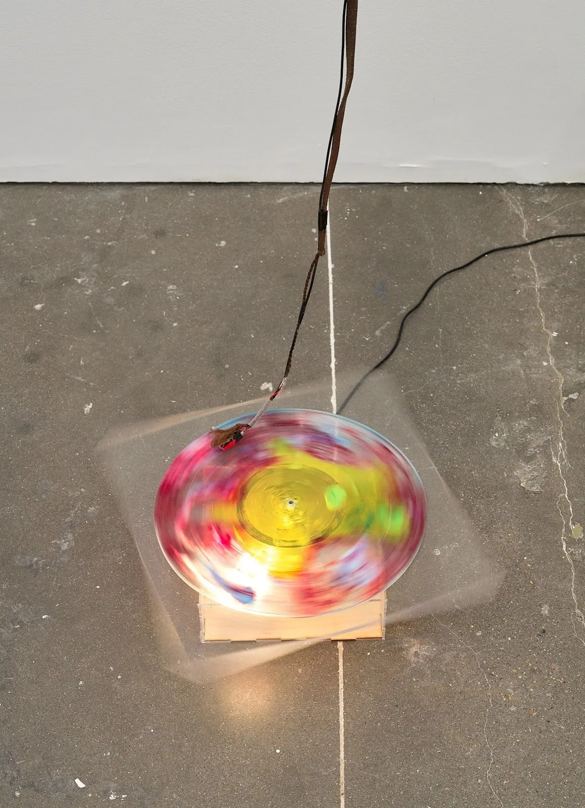 On a concrete floor, a mid-movement clear record player spins a transparent, multicolored vinyl record. Wires come out of the structure and hang from above.