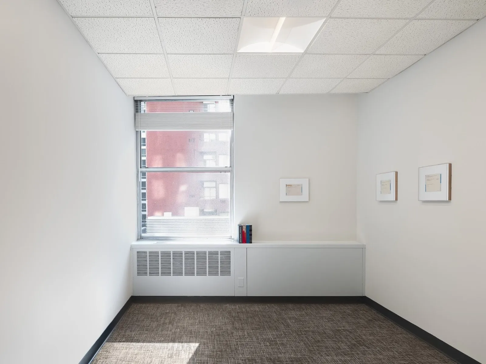 Three framed telegram artworks by On Kawara hang on a pale wall in a bright, modern room with a large window and neutral carpeting.
