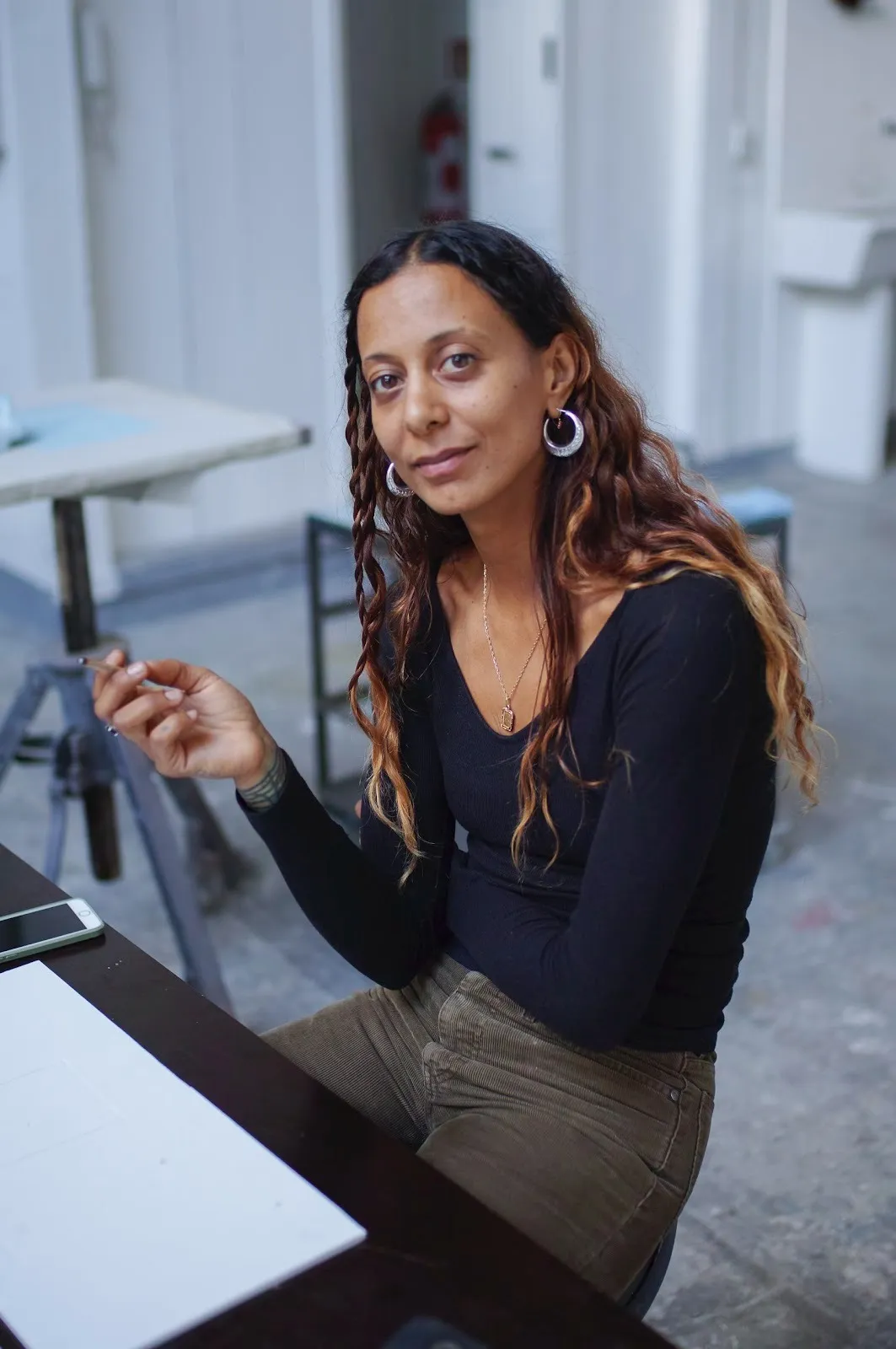 A person in a black tip and grey jeans with long, wavy brown hair with blonde ends sits at a table and faces the camera, smiling.