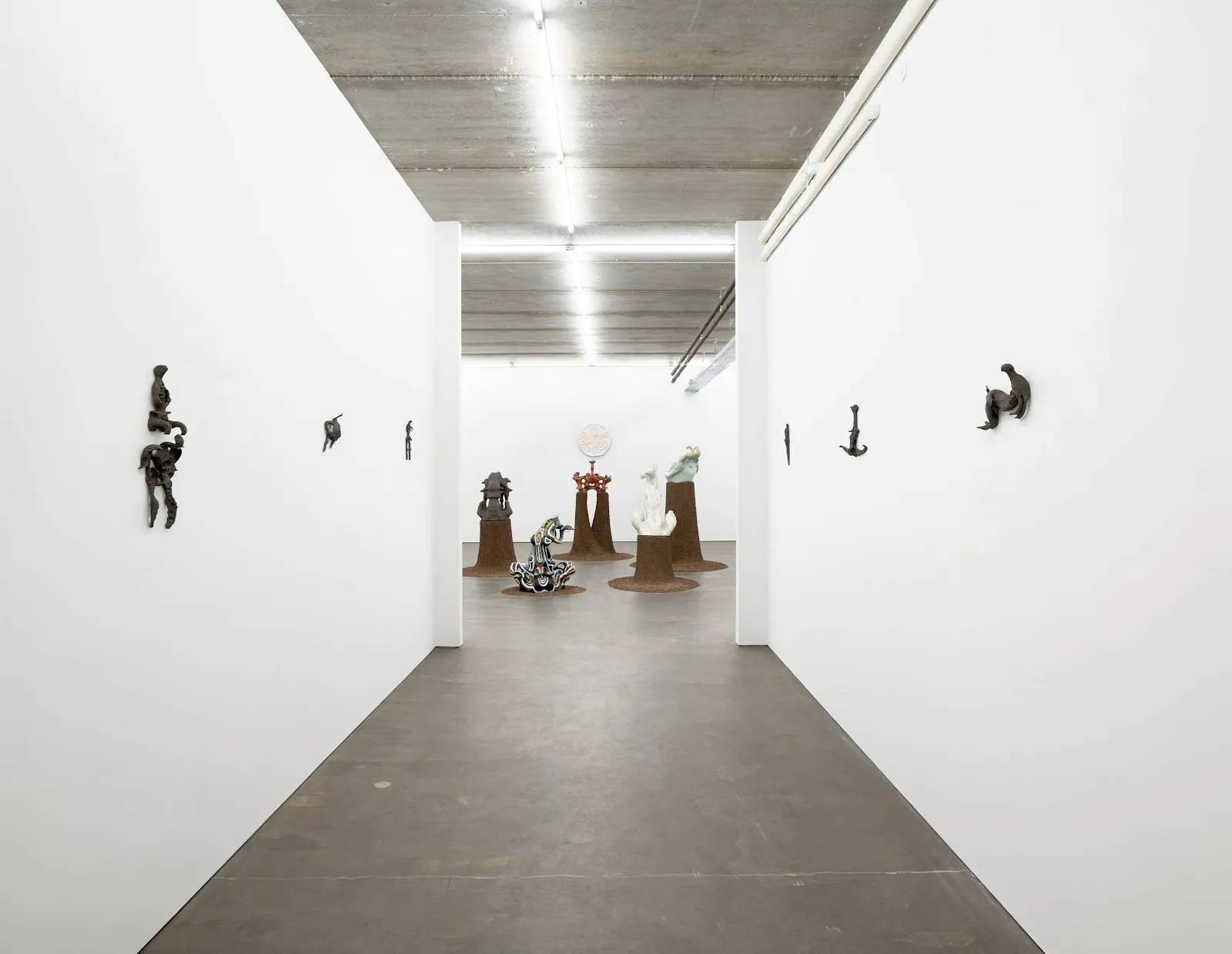 A white-walled gallery corridor, lined sparsely with small, black wall-mounted sculptures, leads into a larger room, where different sculptures of size and color sit on circular brown plinths, gathered in one spot.