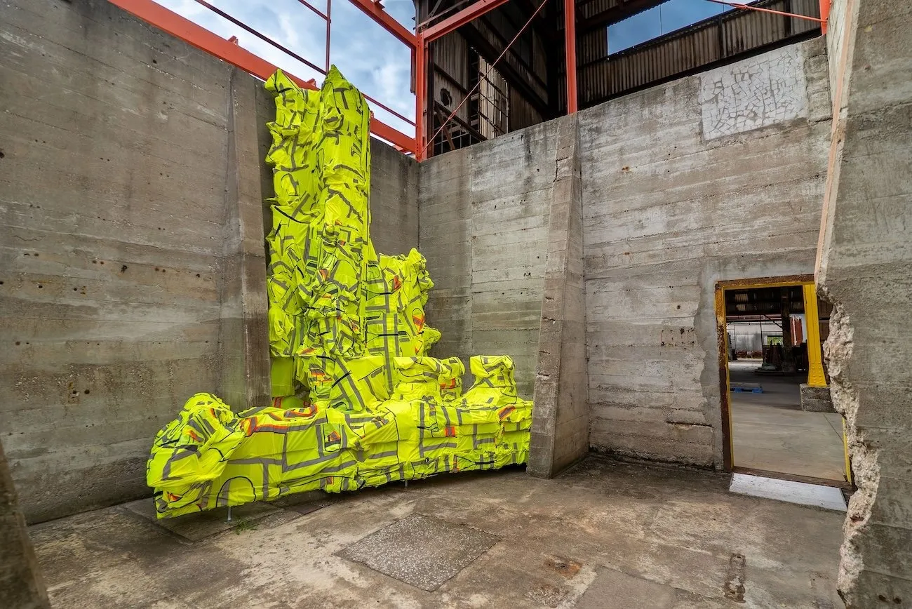 A concrete warehouse structure features a large sculpture that takes up the entire corner. The sculpture has a geometric, lopsided shape covered by neon green construction vest-like fabric.